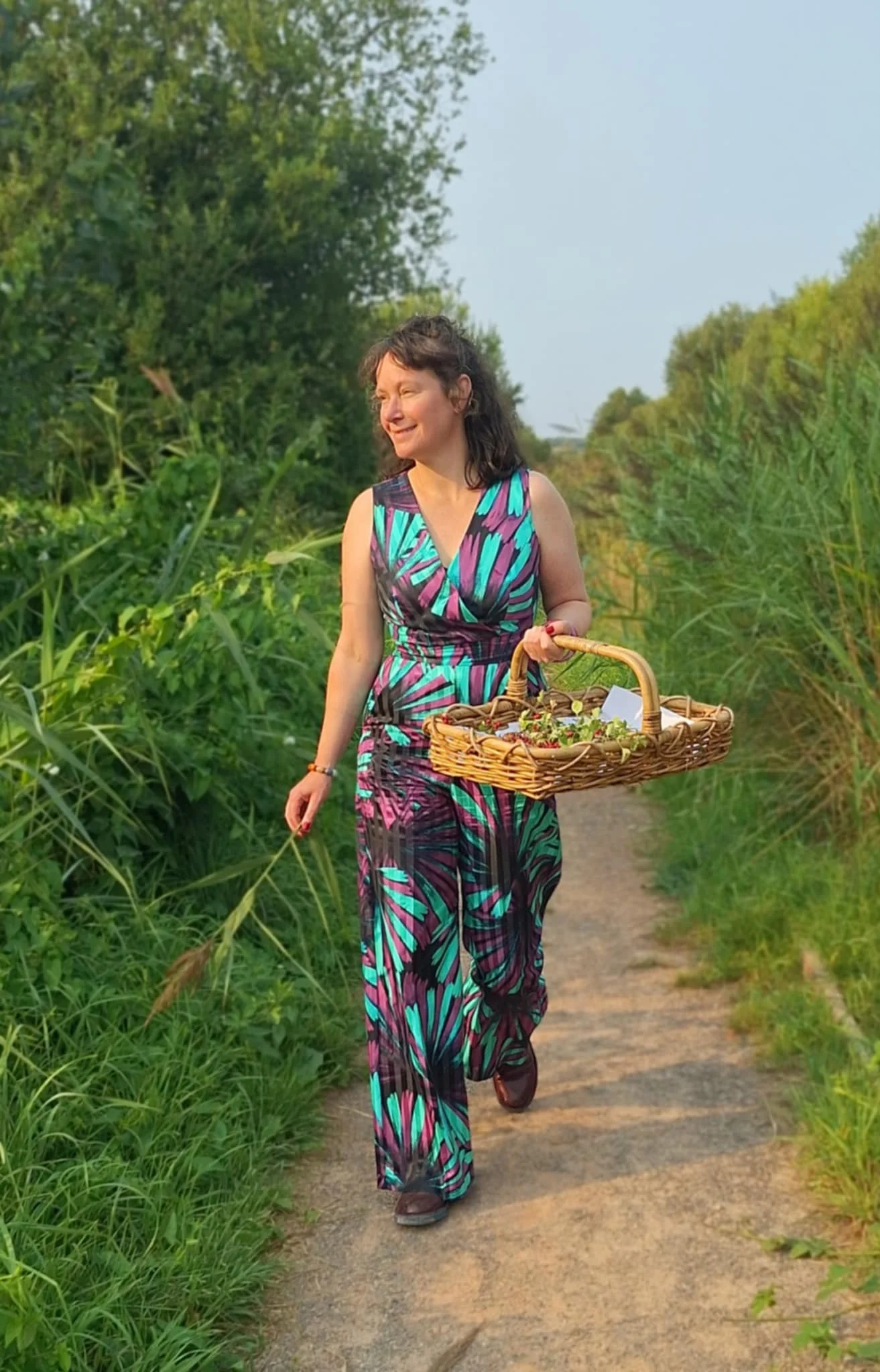 A woman walking along a dirt path surrounded by tall green grass and trees, carrying a woven basket filled with herbs.