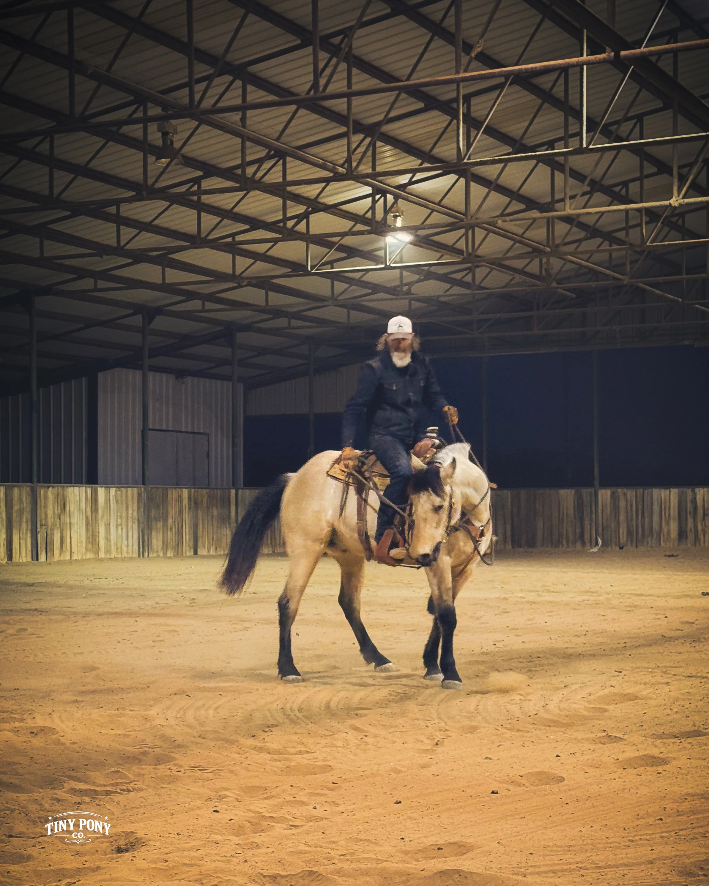 A person riding a horse inside an indoor riding arena at night. The rider wears a hat and dark clothing, and the arena has a dirt floor, metal walls, and a high ceiling with metal beams and lights.