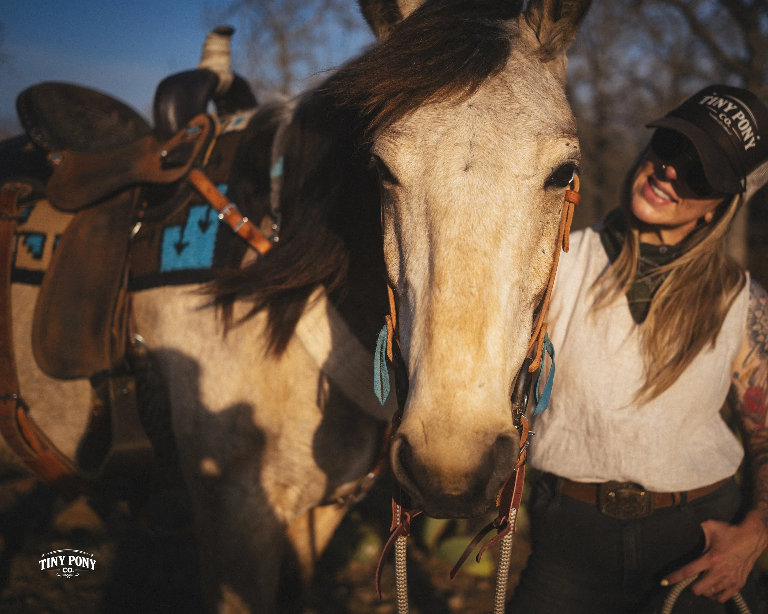 A woman smiling beside a brown and white horse with a saddle, wearing a black cap and sunglasses, during sunset.