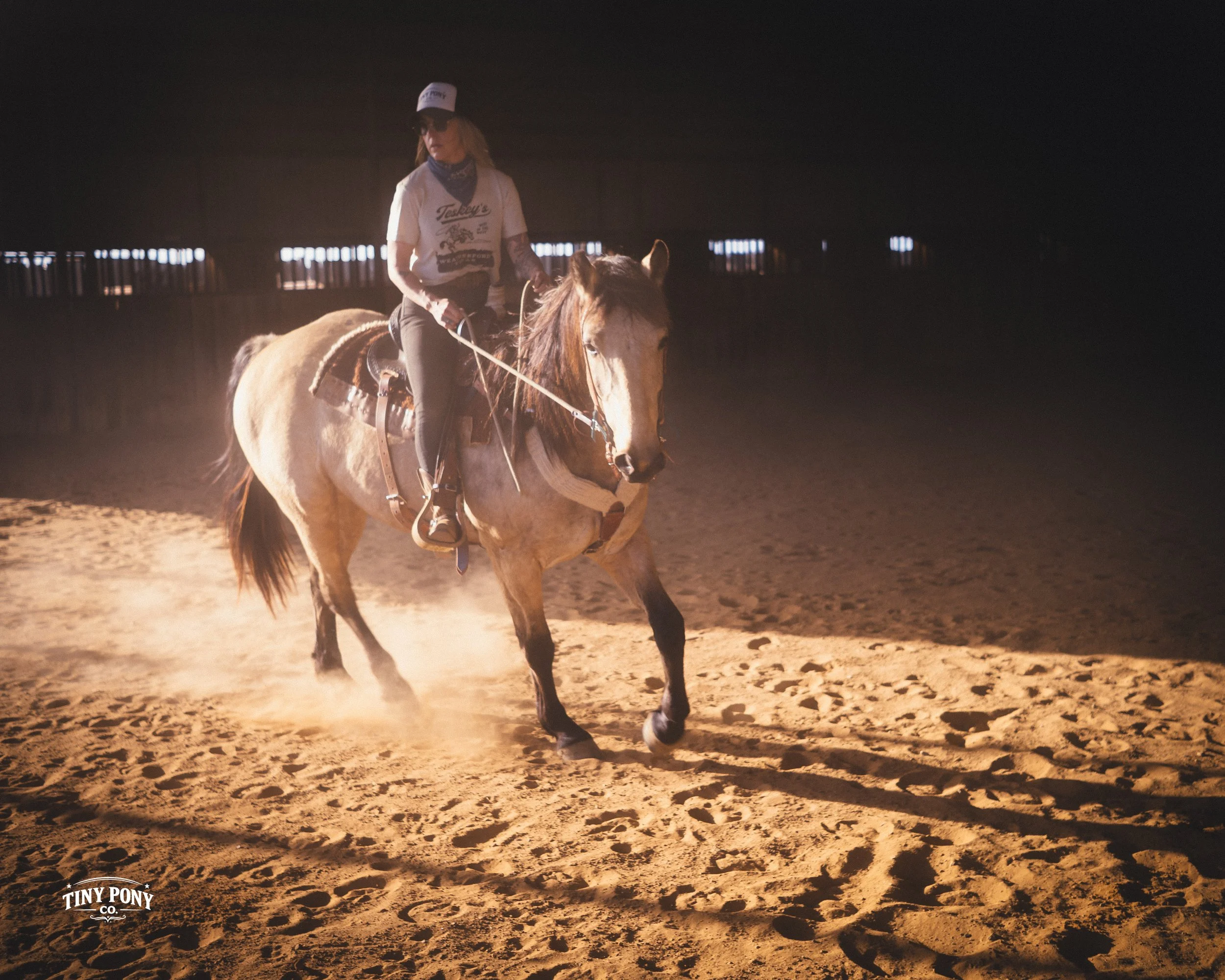 A woman riding a horse in an indoor riding arena at night, with dust kicking up from the sandy ground.