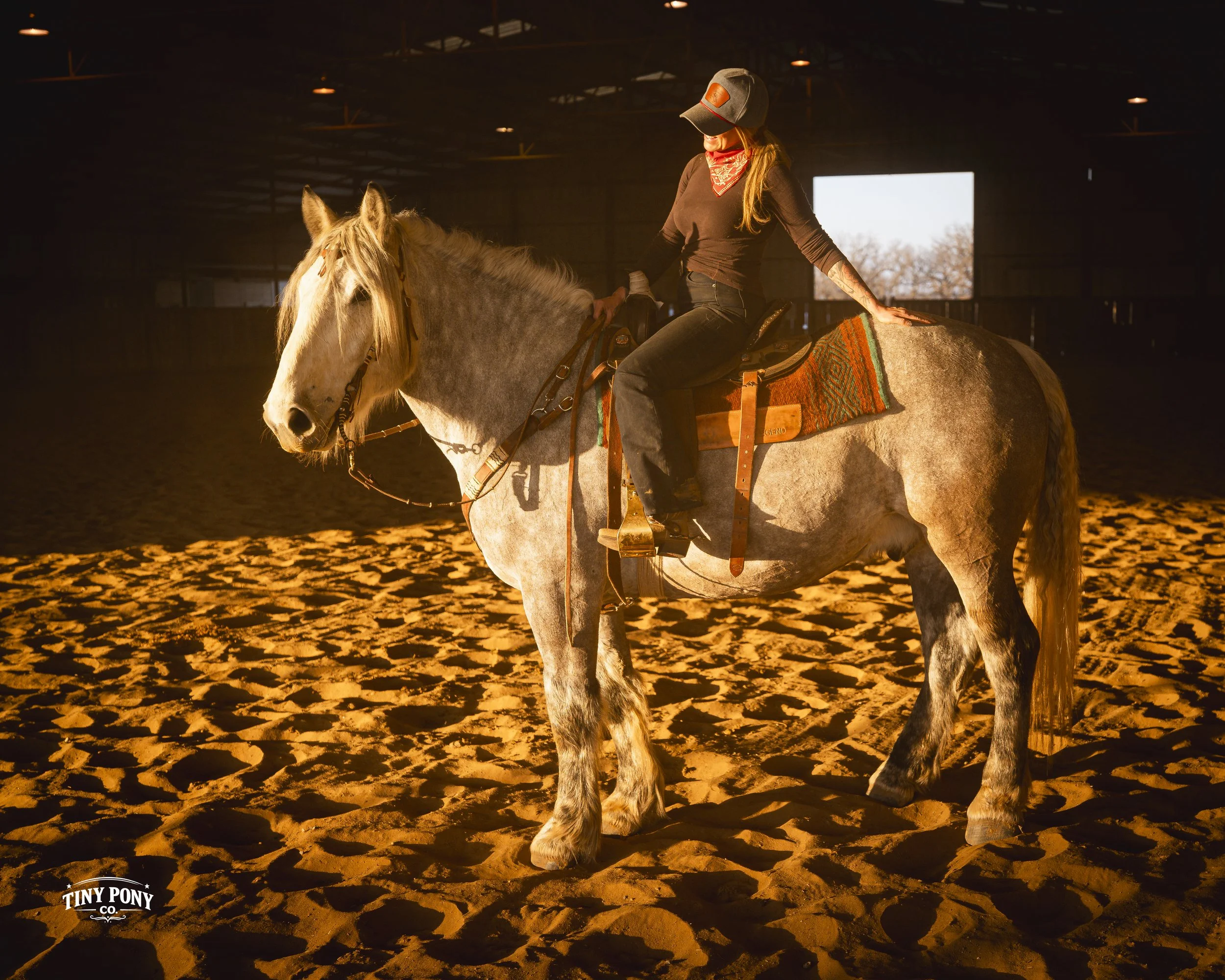 A woman wearing a hat, red bandana, and black long-sleeve shirt sits on a white and gray horse inside an indoor riding arena with sandy ground, dim lighting, and a large open window in the background.
