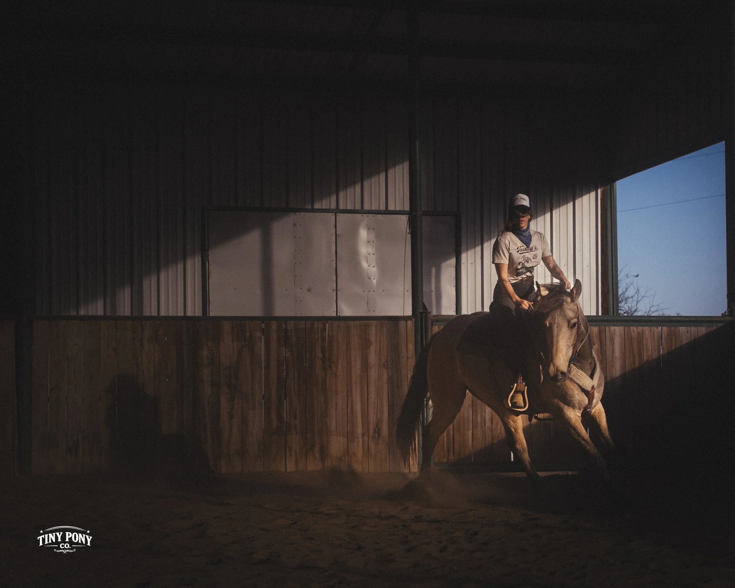 A woman riding a horse in an indoor riding arena with sunlight casting shadows on the wooden walls and dirt floor. She is wearing a cap, sunglasses, and casual riding clothes.