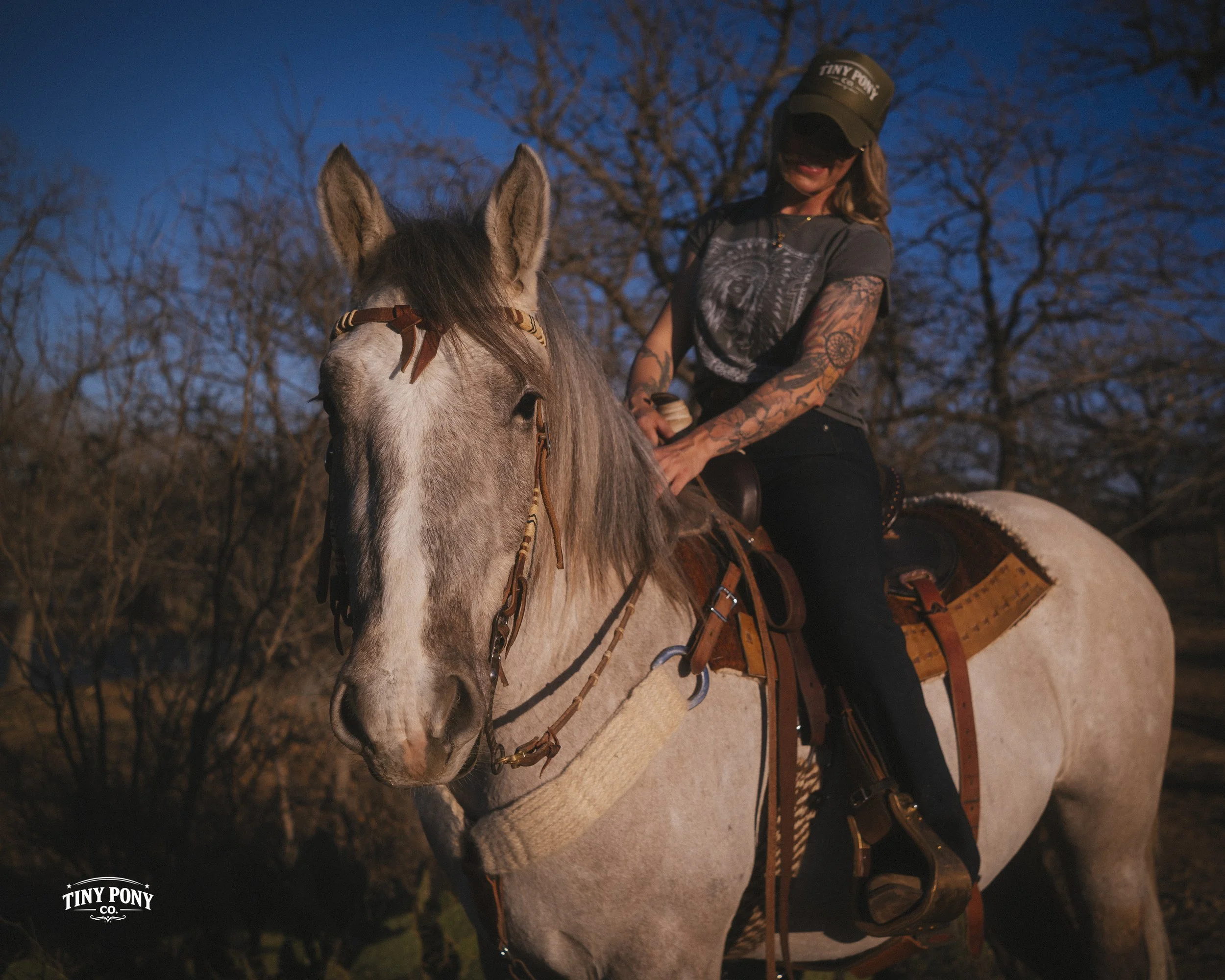 A woman with tattoos wearing a cap and black pants is riding a light gray horse during dusk or dawn, with leafless trees in the background.