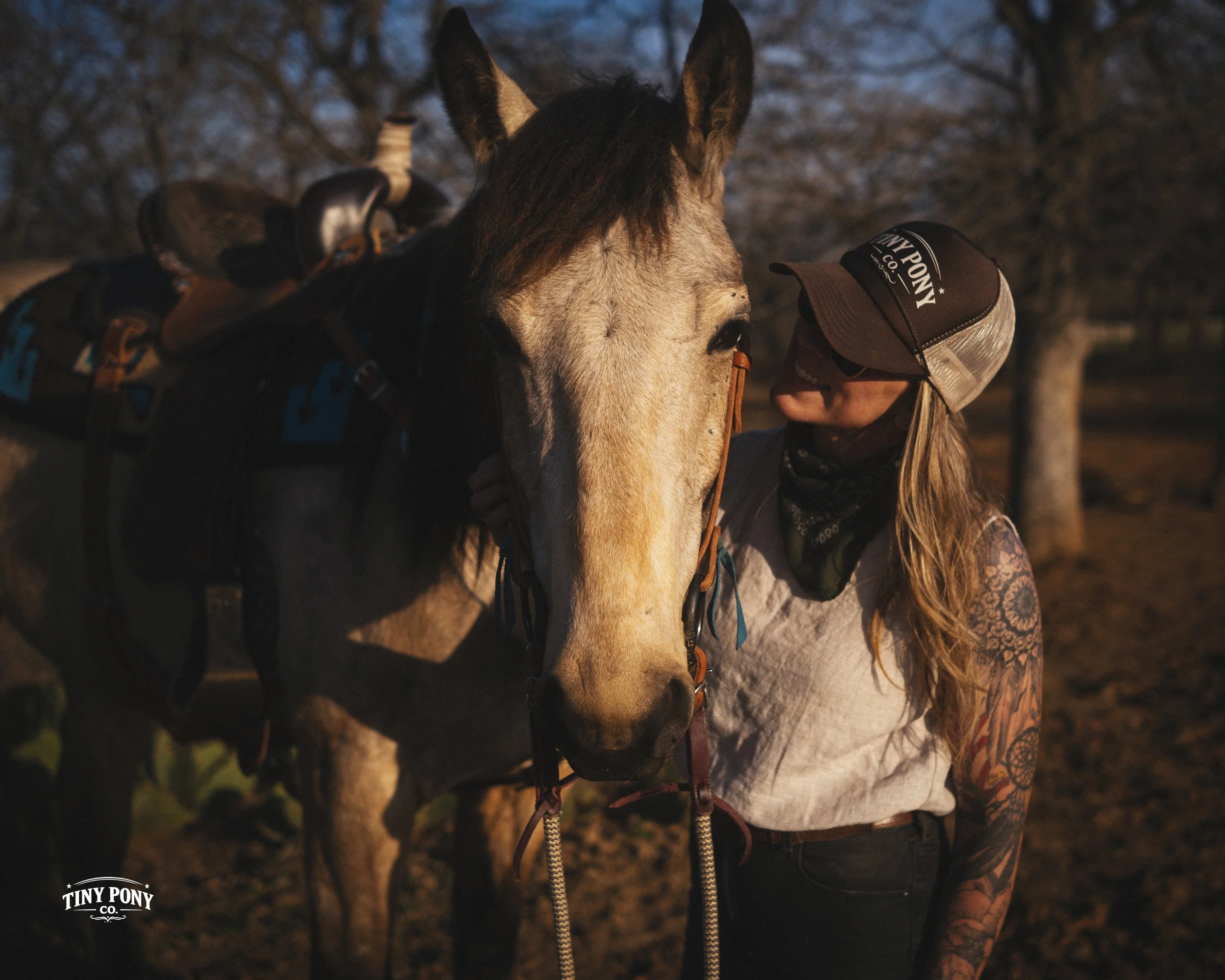 A woman with tattoos on her arm wearing a cap, glasses, and bandana, is standing close to a horse, gently touching its face in an outdoor setting during sunset or sunrise.