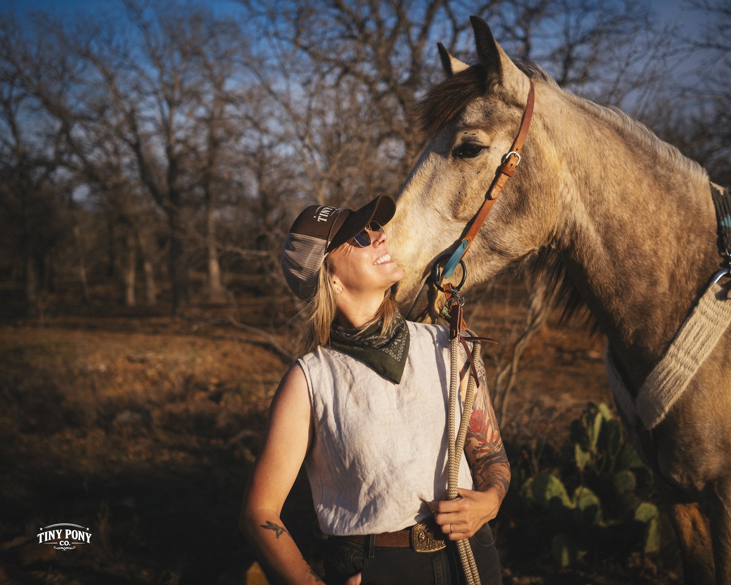 A woman with tattoos, wearing sunglasses, a cap, and a bandana, stands outdoors holding a lead rope, smiling as a horse nuzzles her face in a desert landscape with leafless trees.