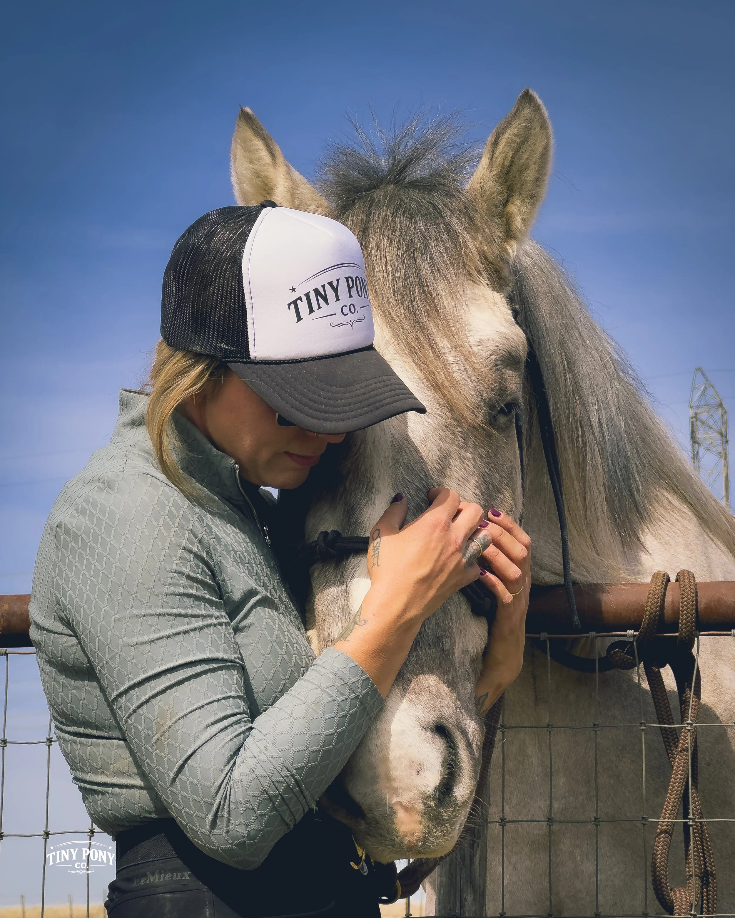 A woman wearing a gray jacket and a white and black trucker hat with 'Tiny Pony Co.' written on it is hugging a light gray and white horse through a metal fence. The horse is leaning its head down, and the woman is gently holding its face.