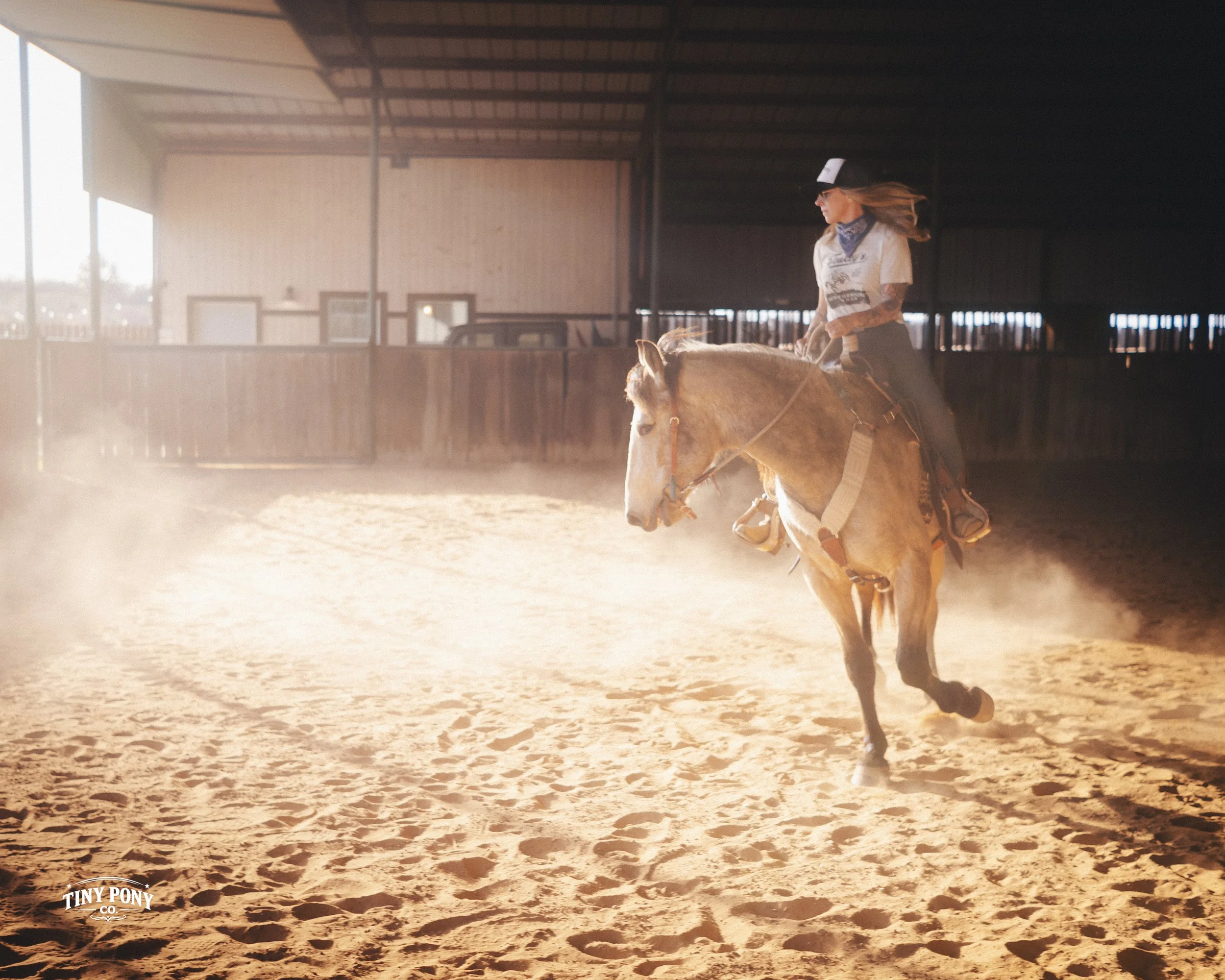A woman riding a horse inside an indoor riding arena during daytime with sunlight streaming in, kicking up dust as the horse moves.