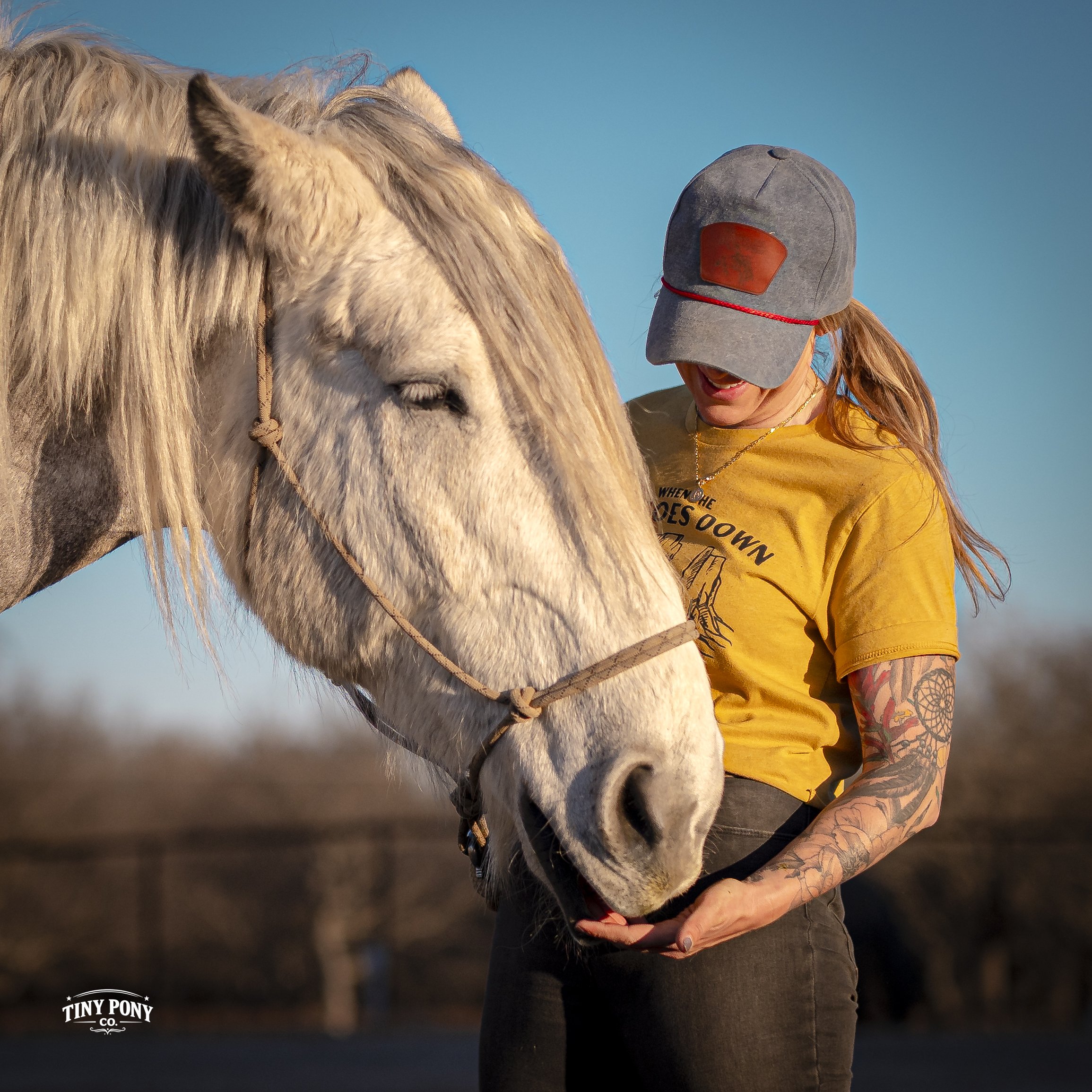 A woman with tattoos on her arm wearing a gray cap and yellow t-shirt, gently holding a white horse's chin with her hands during the daytime.