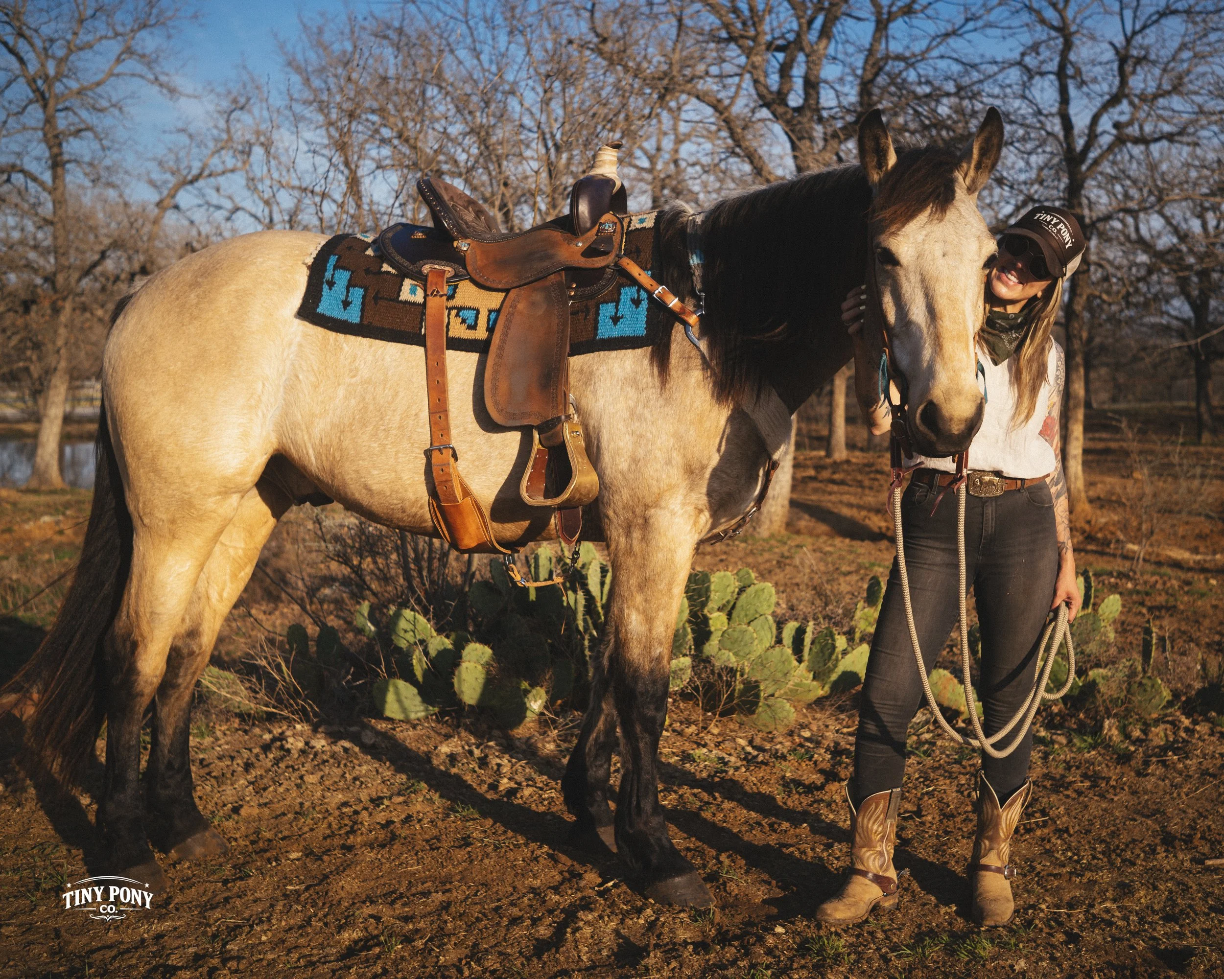 A woman wearing sunglasses, a cap, and cowboy boots smiling and petting a light-colored horse with a dark mane, in a rural outdoor setting with leafless trees and cacti.