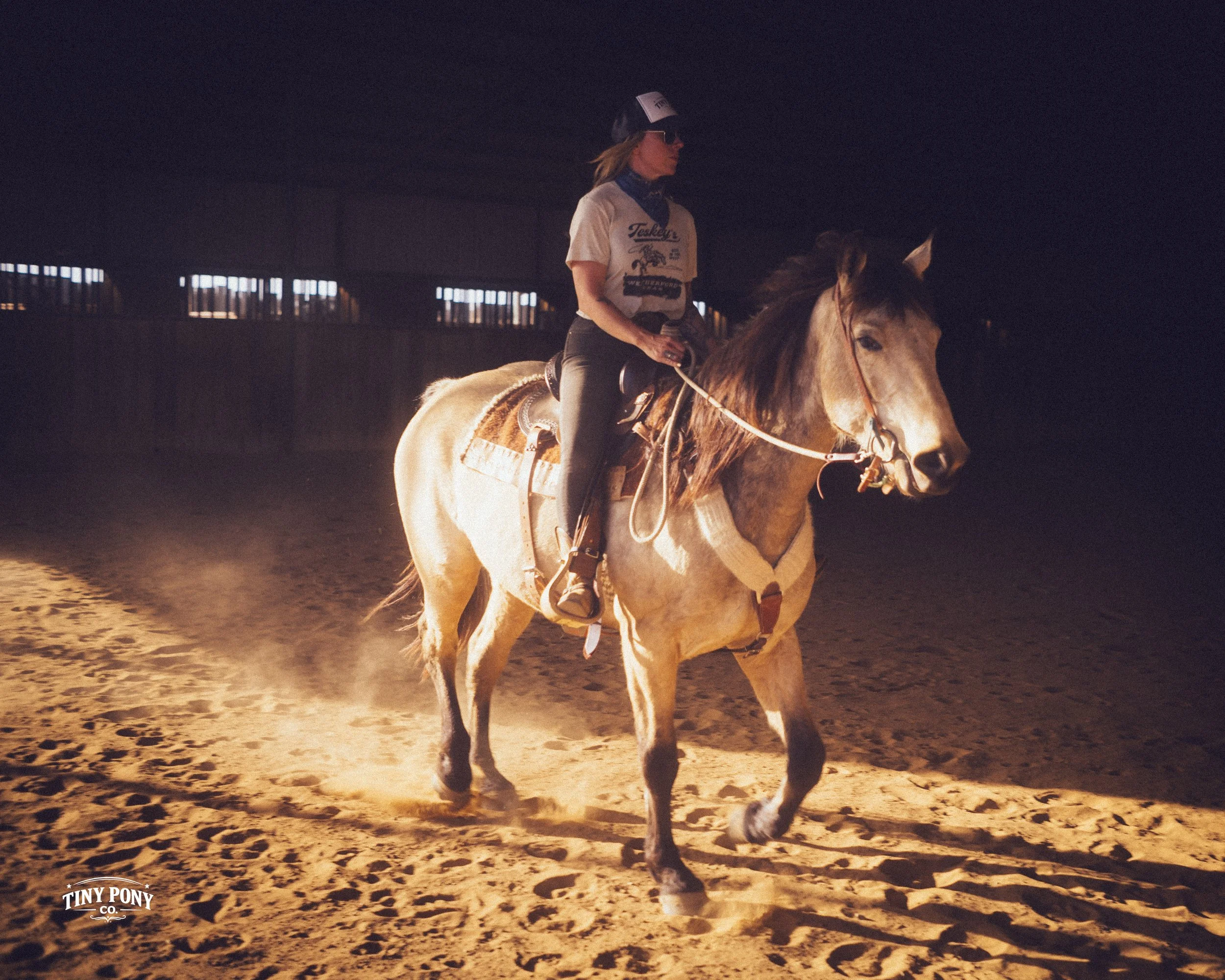 A woman riding a horse in a dimly lit indoor arena, illuminated by a single light source, with a dark background and sandy floor.