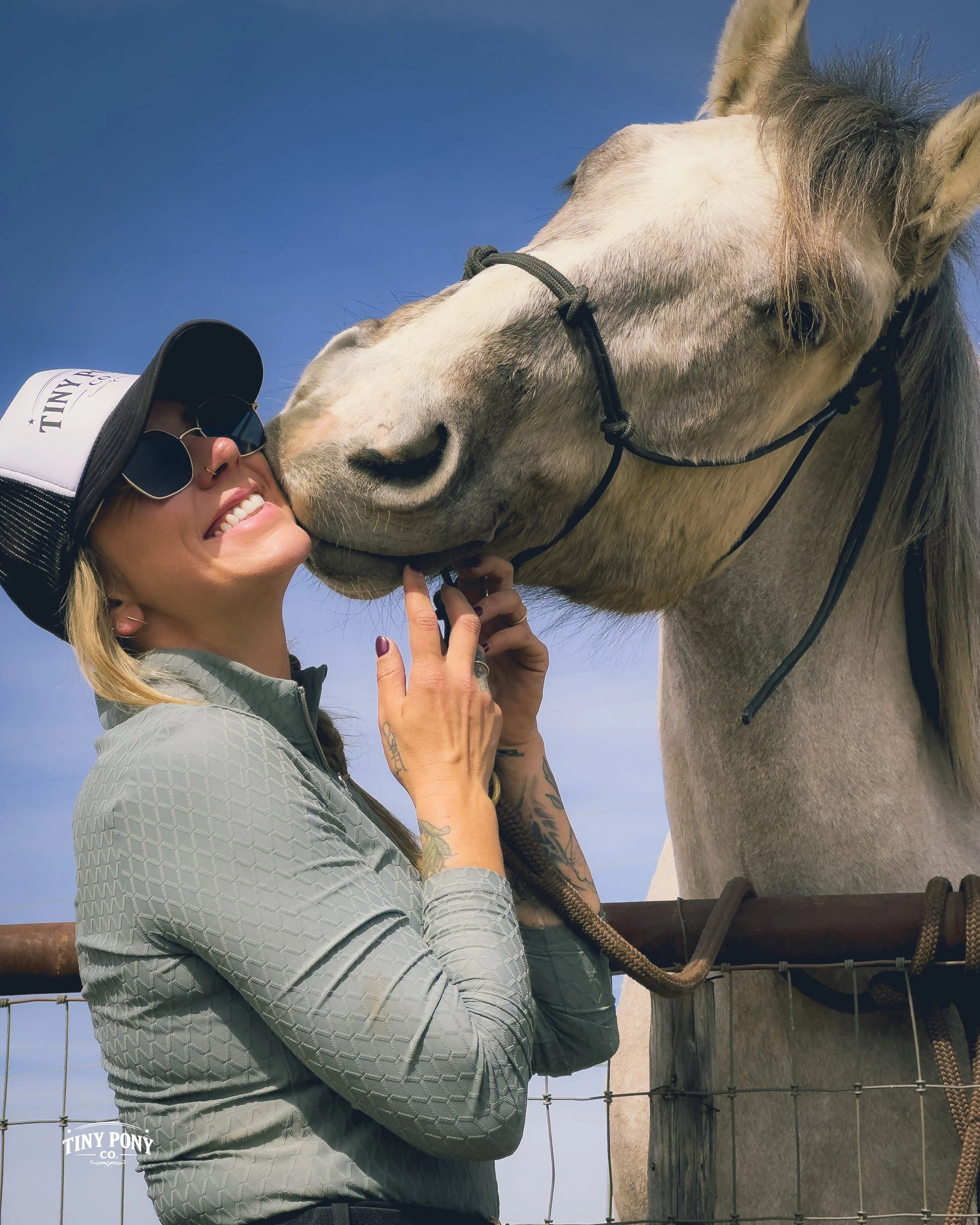 Woman smiling and hugging a white horse with a gray mane, wearing sunglasses and a gray jacket, outdoors against a blue sky