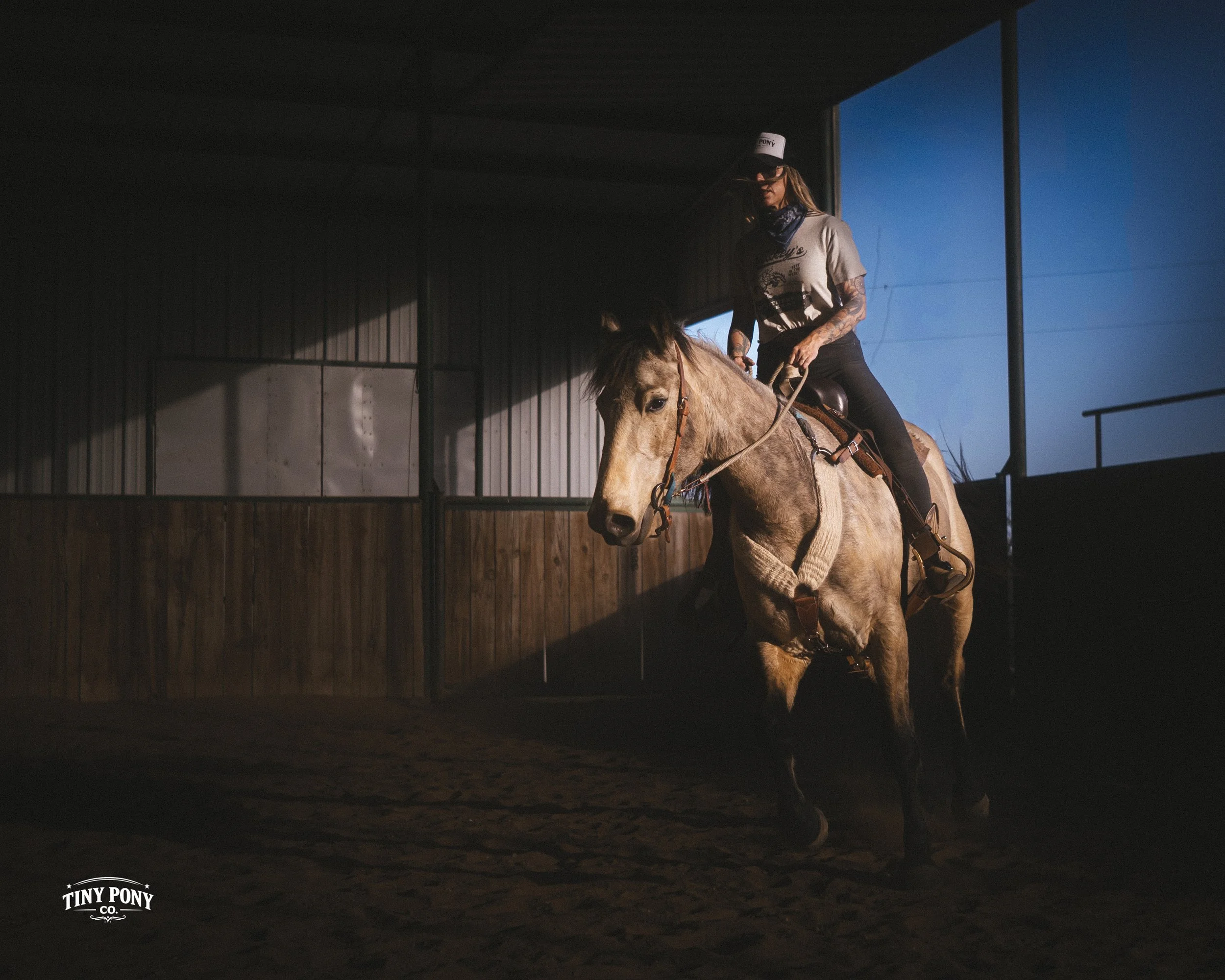 A woman riding a light brown horse inside an indoor riding arena during late afternoon or evening with sunlight streaming through a large open window.