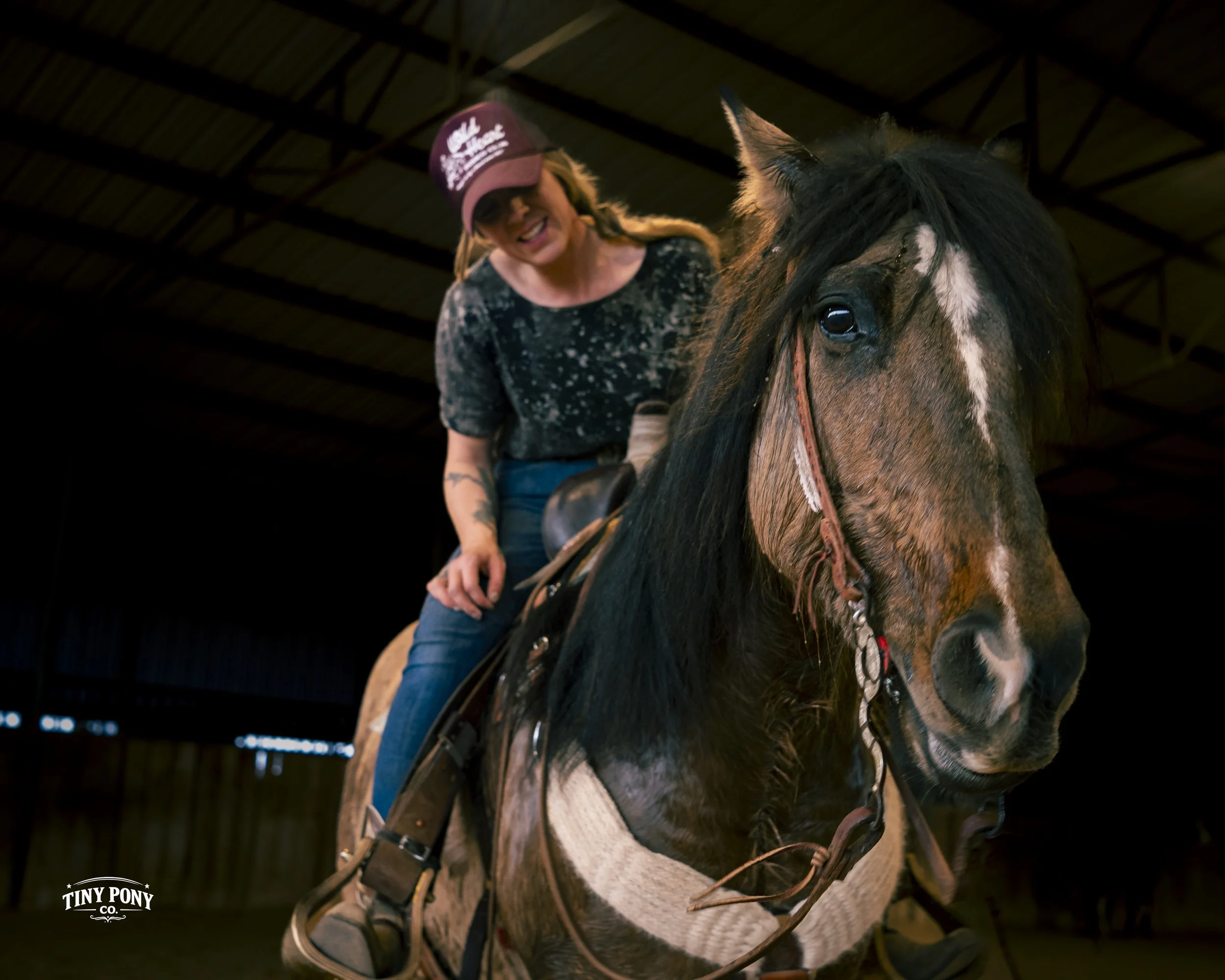 A woman riding a brown and black horse inside a barn. The woman is smiling and wearing a purple cap and a black shirt. The horse is wearing a riding saddle and bridle.