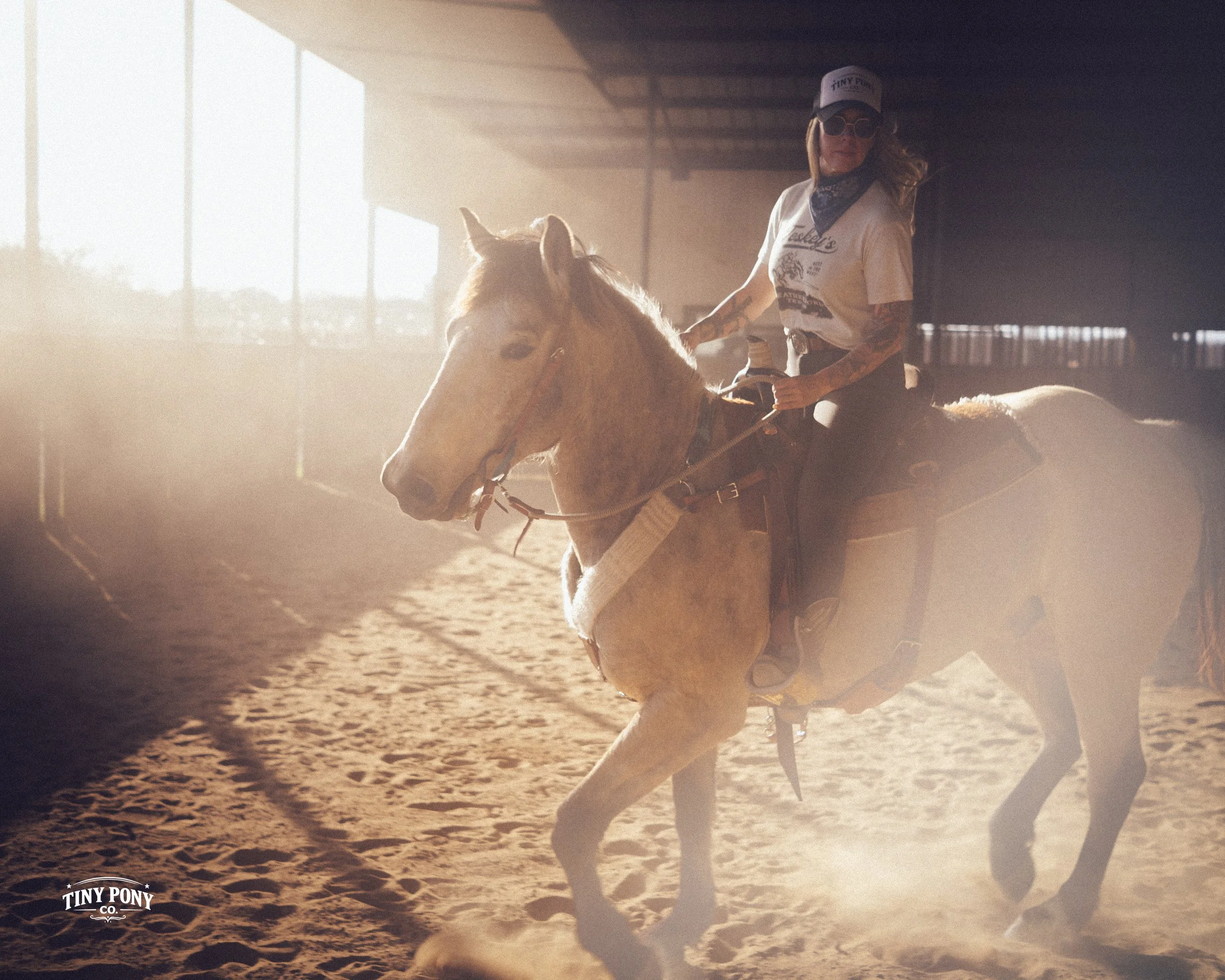 A woman with tattoos, wearing a cap, sunglasses, bandana, and casual shirt, is riding a light-colored horse inside an indoor riding arena with sunlight streaming through large windows.