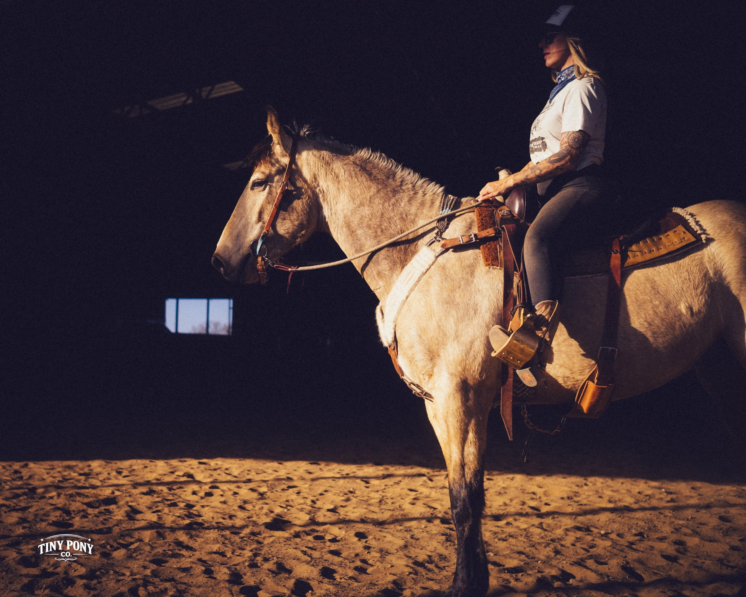 A woman wearing a cowboy hat, sunglasses, and a bandana around her neck riding a light brown horse inside an indoor riding arena with a sandy floor.