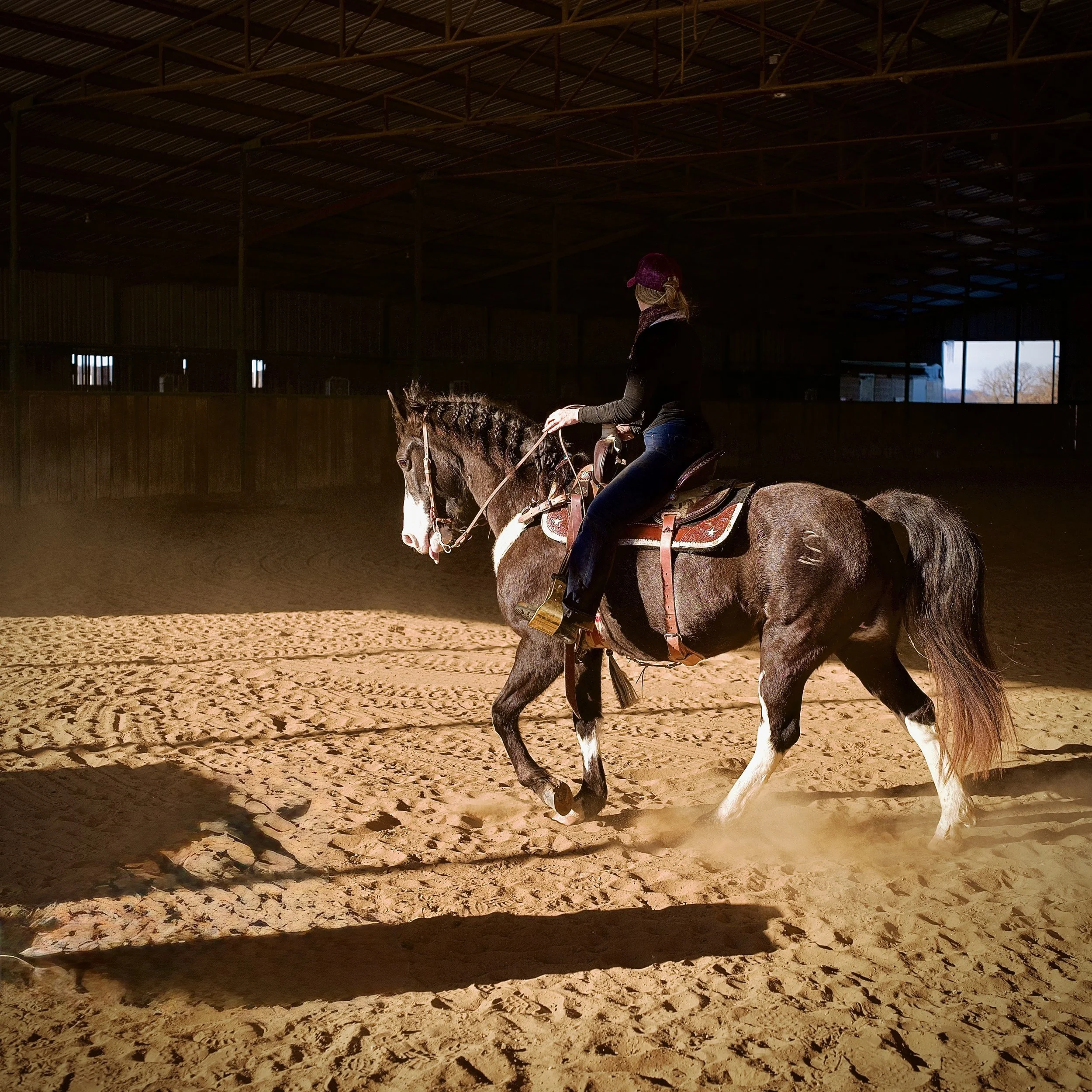 Woman riding a horse in an indoor riding arena with sandy footing and low lighting.