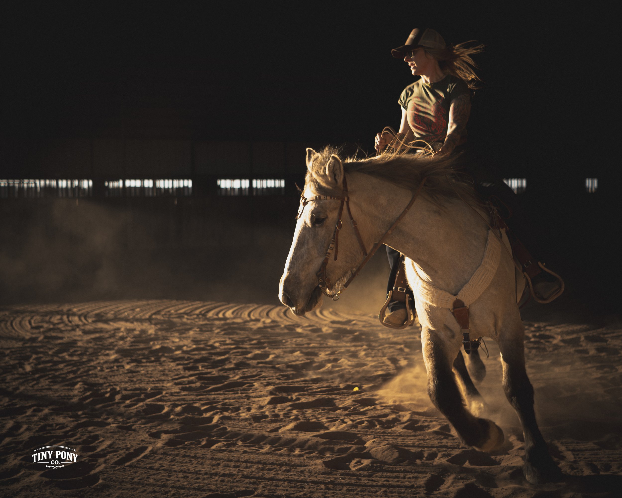 A woman riding a buckskin horse inside an indoor arena with dirt floor, illuminated by natural light from windows