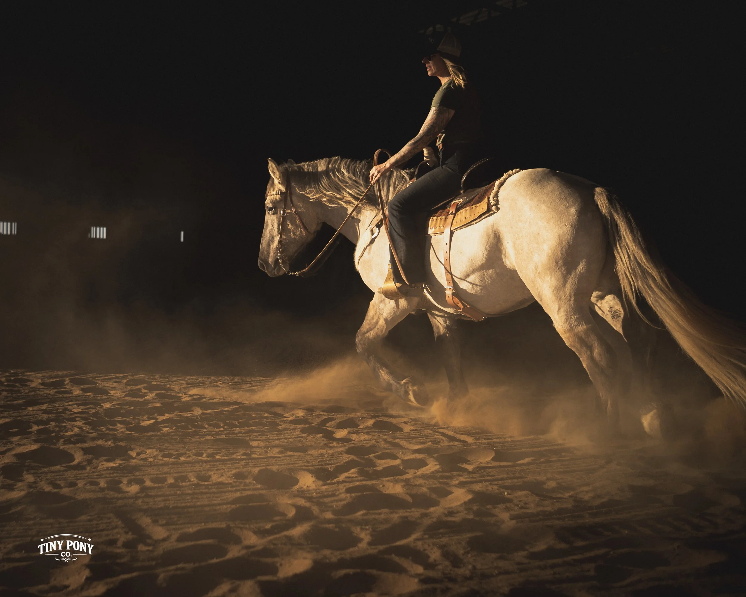 A person riding a white horse at night, kicking up dust on a sandy terrain, with a dark background and minimal lights.