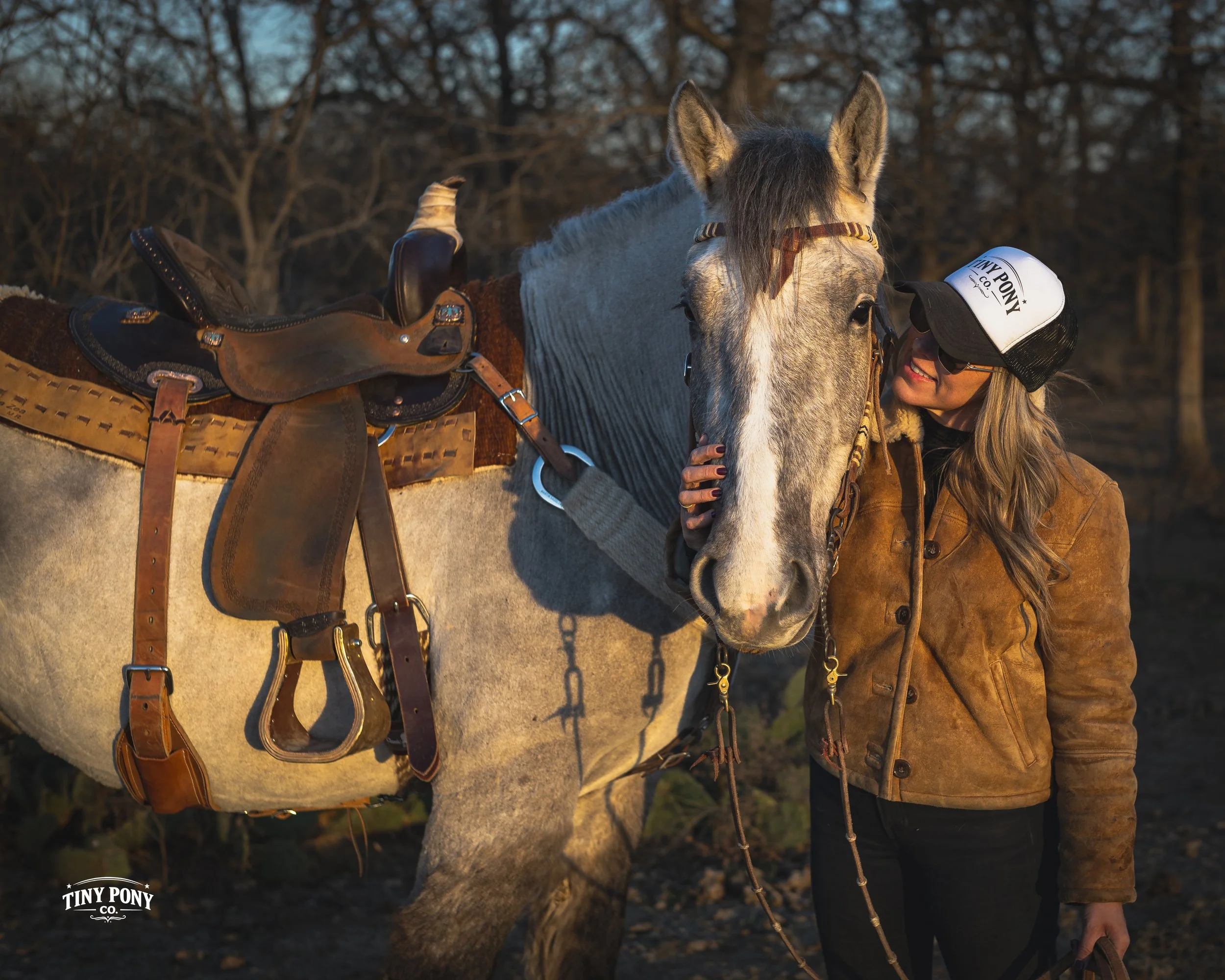 A woman wearing a brown leather jacket and a white cap with 'Tiny Pony Co.' logo, smiling and petting a gray horse with a saddle and bridle, outdoors during late afternoon or early evening.