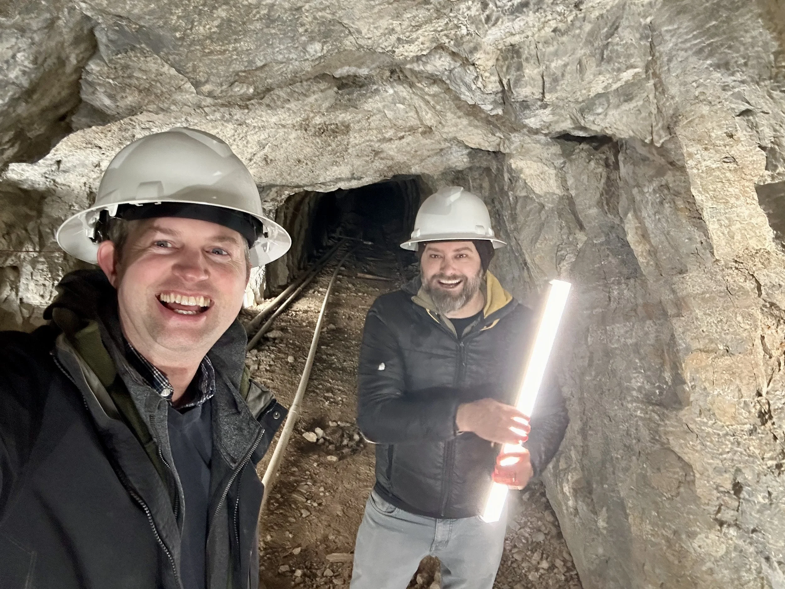 Two men wearing safety helmets inside a mine tunnel, smiling, with one holding a bright light.