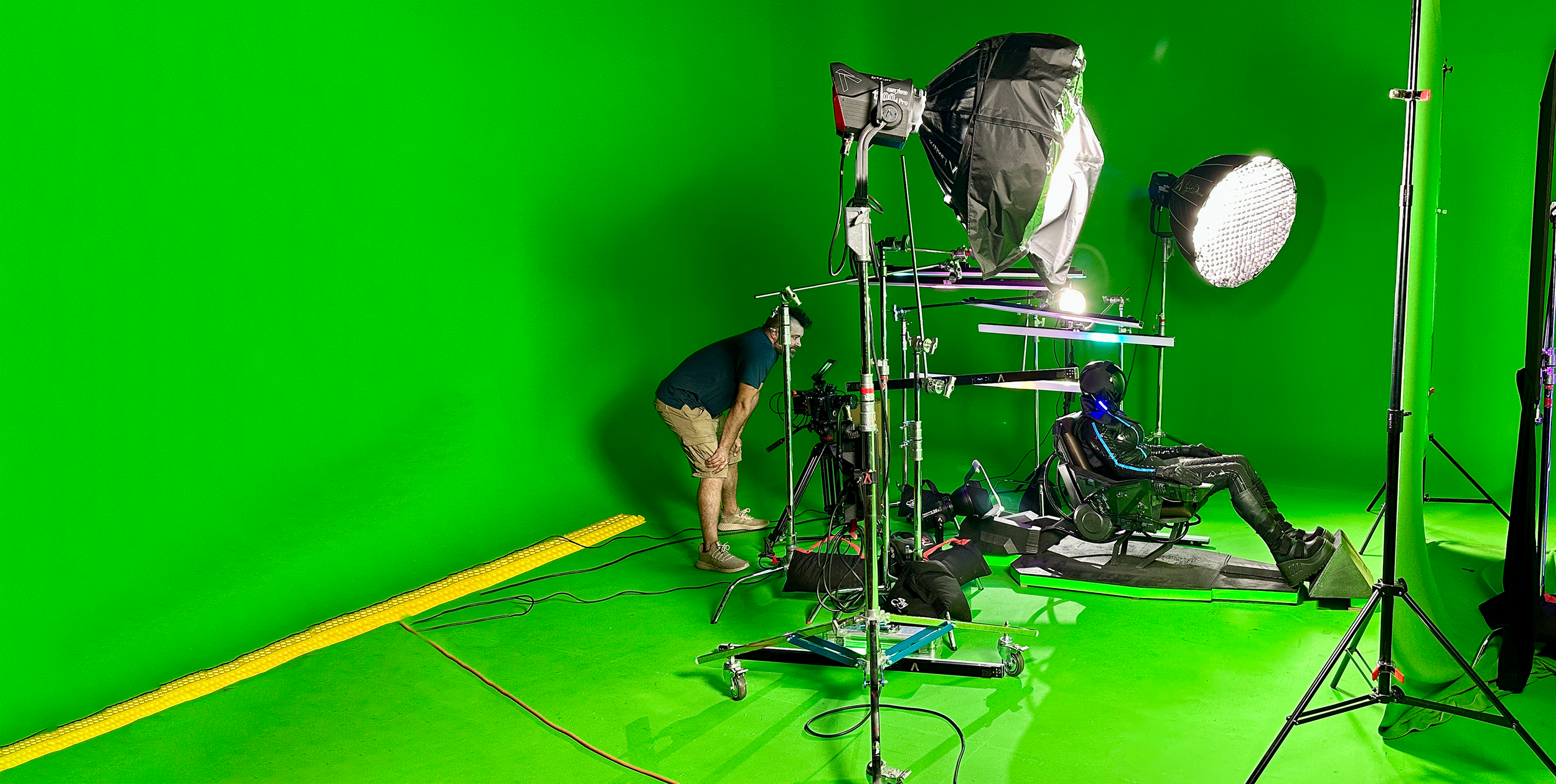 A person sits in front of a green screen during filming, with studio lights and equipment surrounding them.