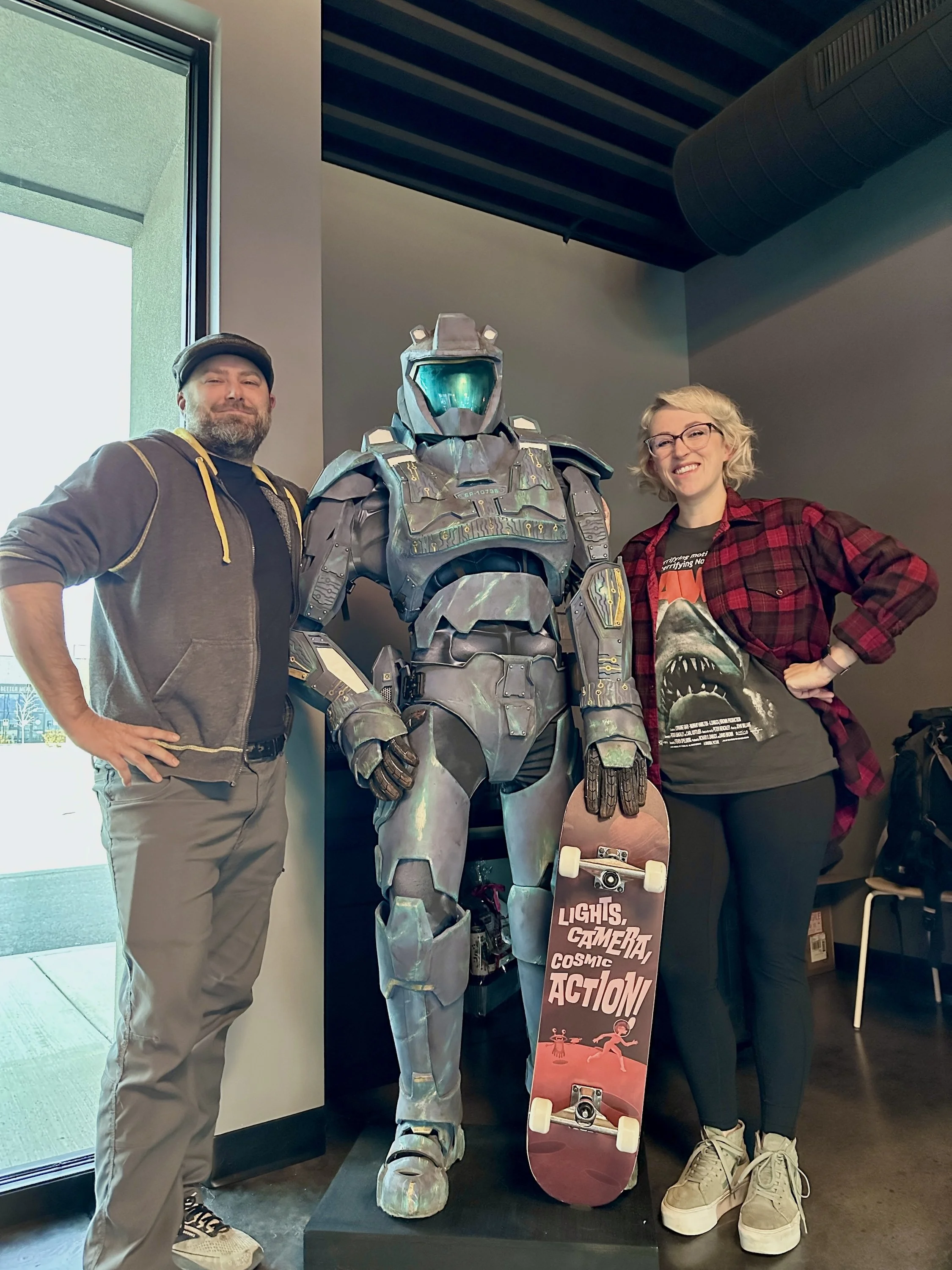 Two people standing next to a life-sized robot sculpture holding a skateboard, inside a modern building with large window and industrial ceiling.