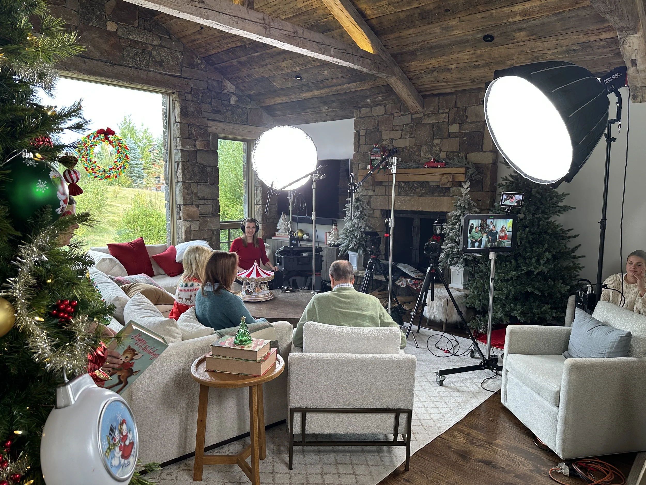 Inside a decorated living room with a Christmas tree, a stone fireplace, and large windows showing a green outdoor view. A small group of people sits on couches and chairs, participating in a holiday video recording or live stream, with professional lighting, cameras, and monitors set up.