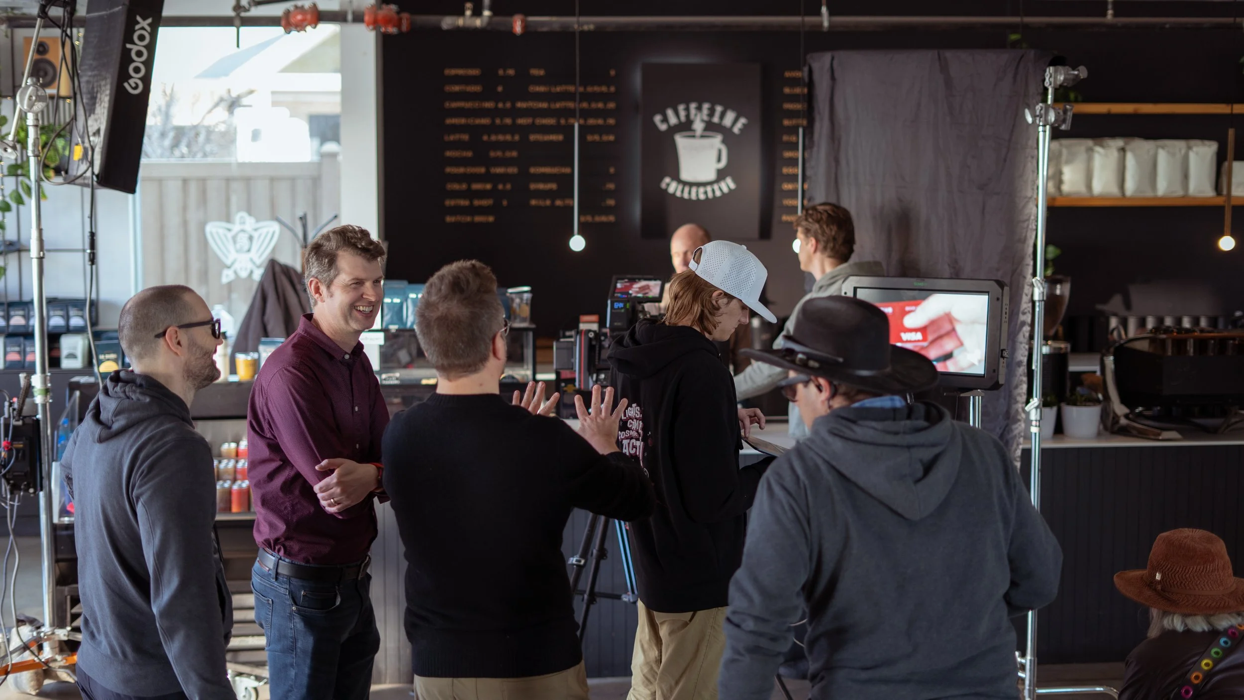 A group of people on set for a filming or photoshoot inside a coffee shop named 'Caffetile Collective.' The team is engaged in conversation and preparing to film or photograph, with various camera equipment and lighting around them.