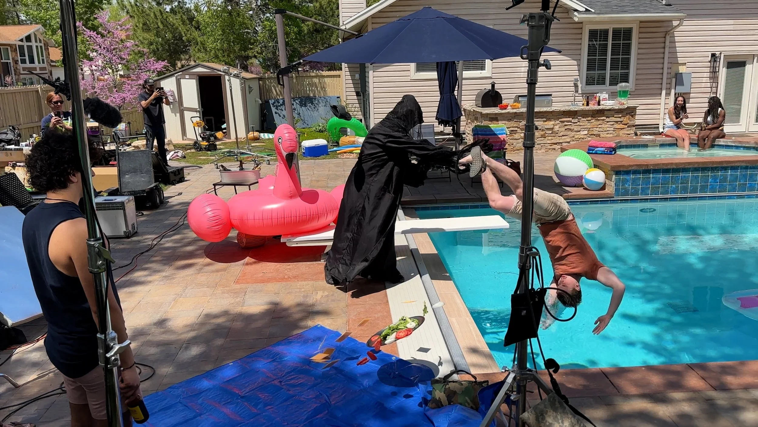 Behind the scenes of a pool party filming, with a person dressed as the grim reaper pushing another person into the pool, a pink flamingo float, people sitting by the pool, and filming equipment.