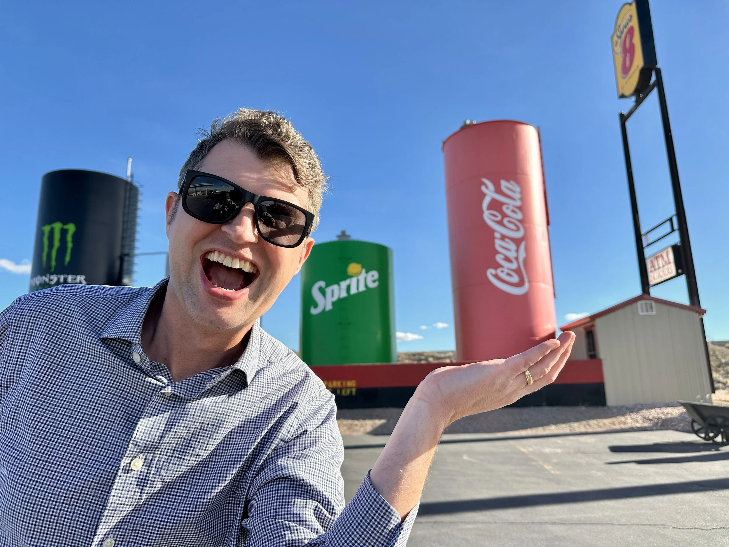 A man wearing sunglasses and a checkered shirt smiling and holding out his hand toward oversized soda cans in the background, including Sprite, Coca-Cola, and Monster, in a parking lot under a blue sky.