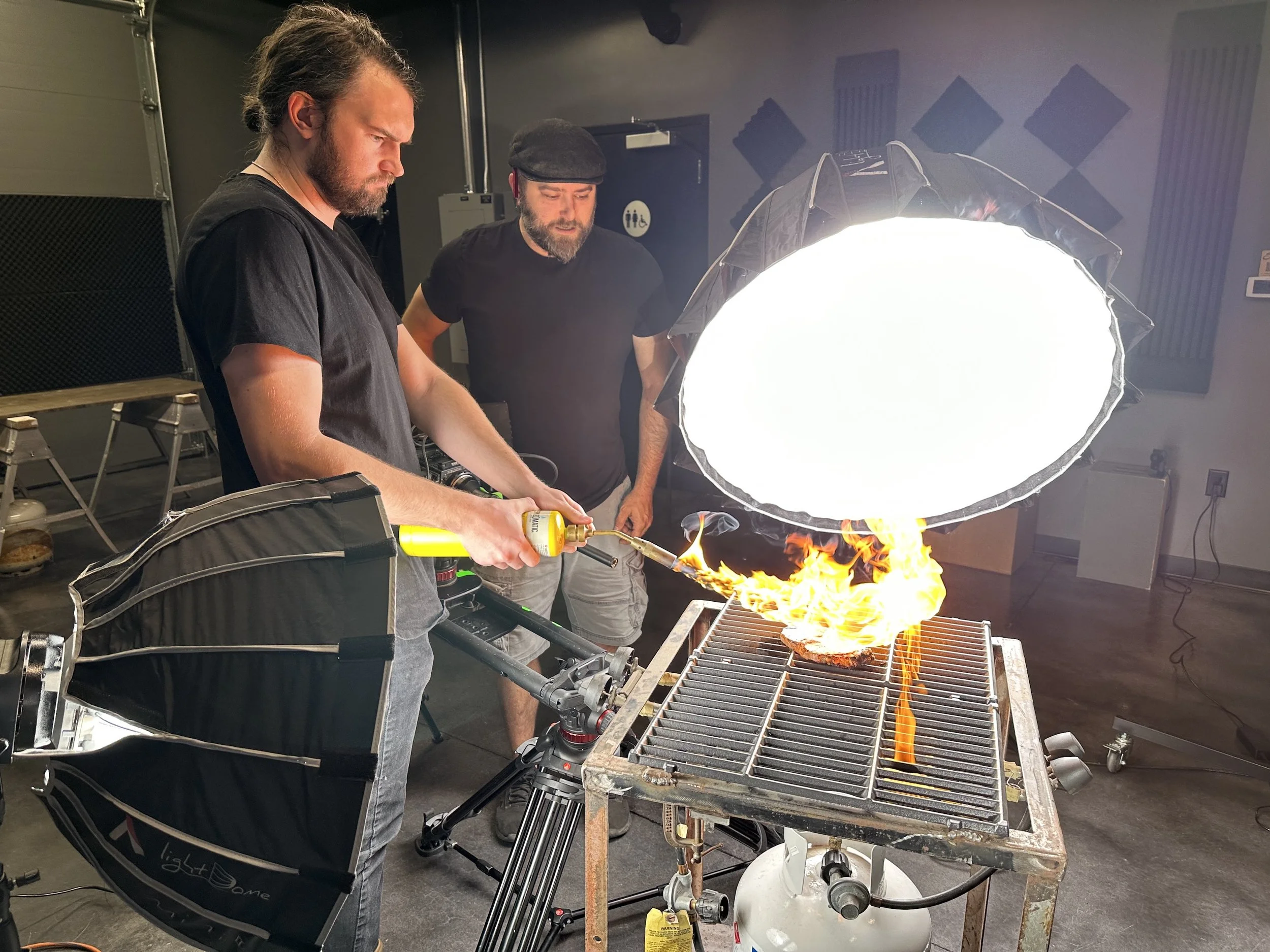 Two men are conducting a photo shoot of a flaming piece of meat on a grill in a studio with professional lighting equipment.