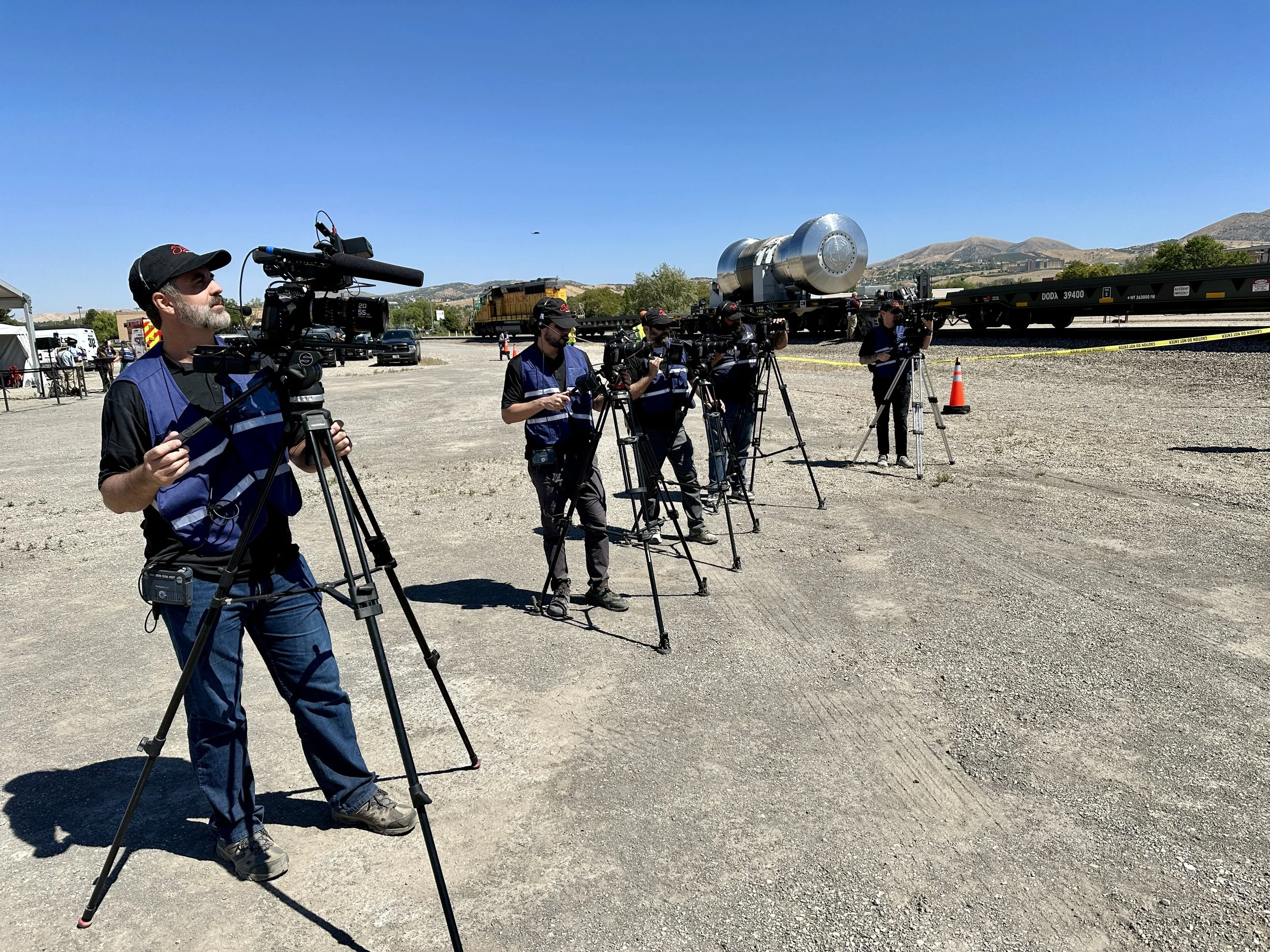 Group of camera operators filming a train with a large silver tank car, set outdoors on a sunny day with mountains in the background.