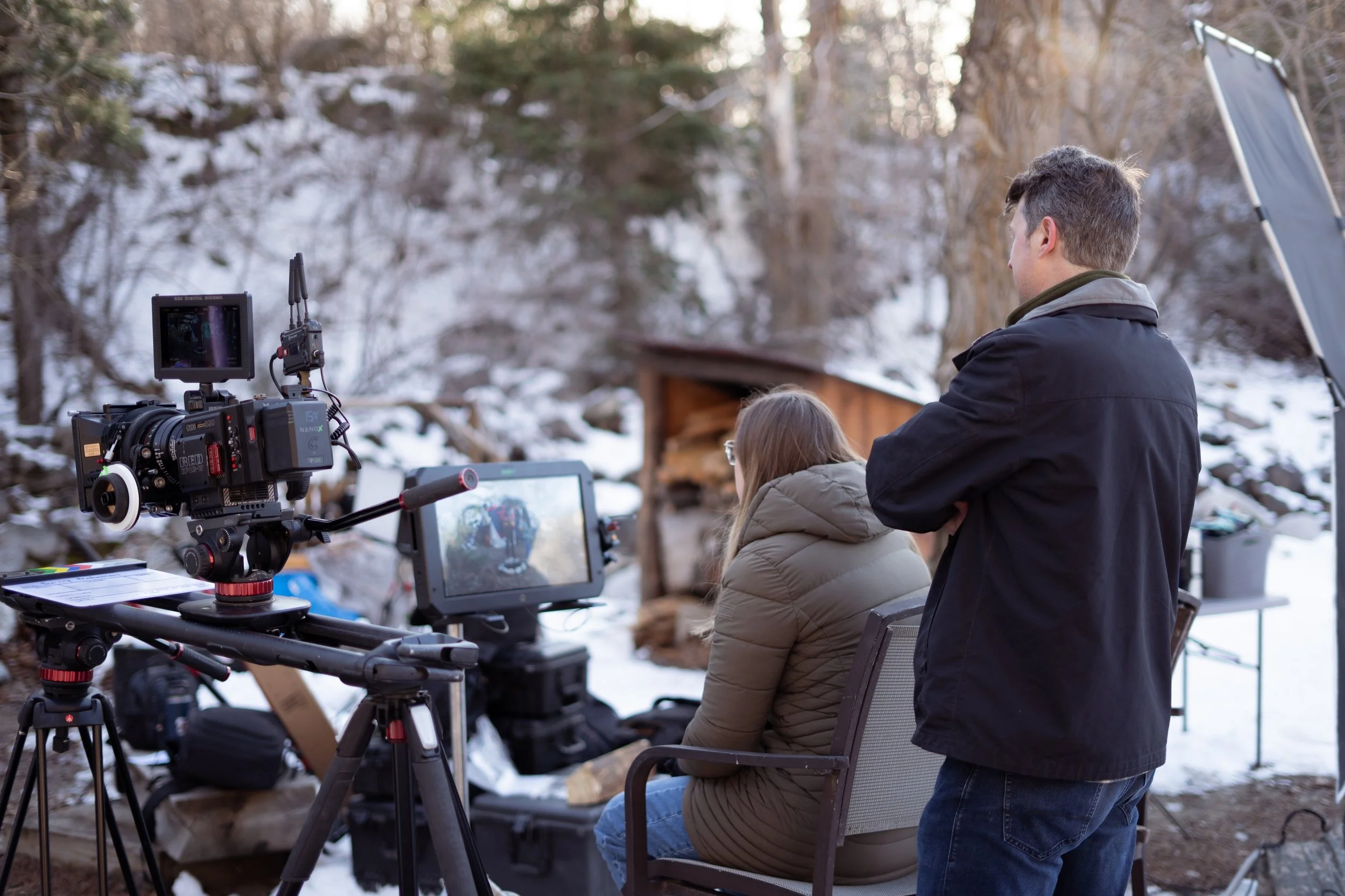Film crew filming outdoors in a snowy, wooded area, with a camera on a tripod facing a woman seated on a chair, and a man standing beside her.