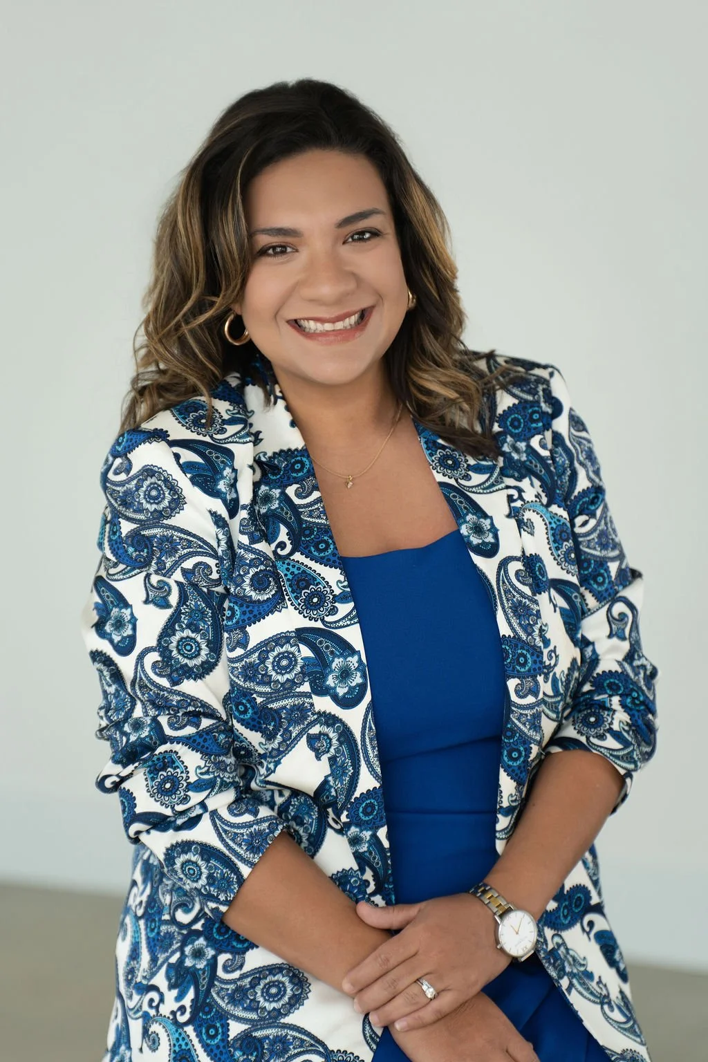 A woman with shoulder-length wavy brown hair, smiling, wearing a blue dress and a white blazer with blue floral patterns, accessorized with gold hoop earrings, a watch, and a ring, posed against a light background.