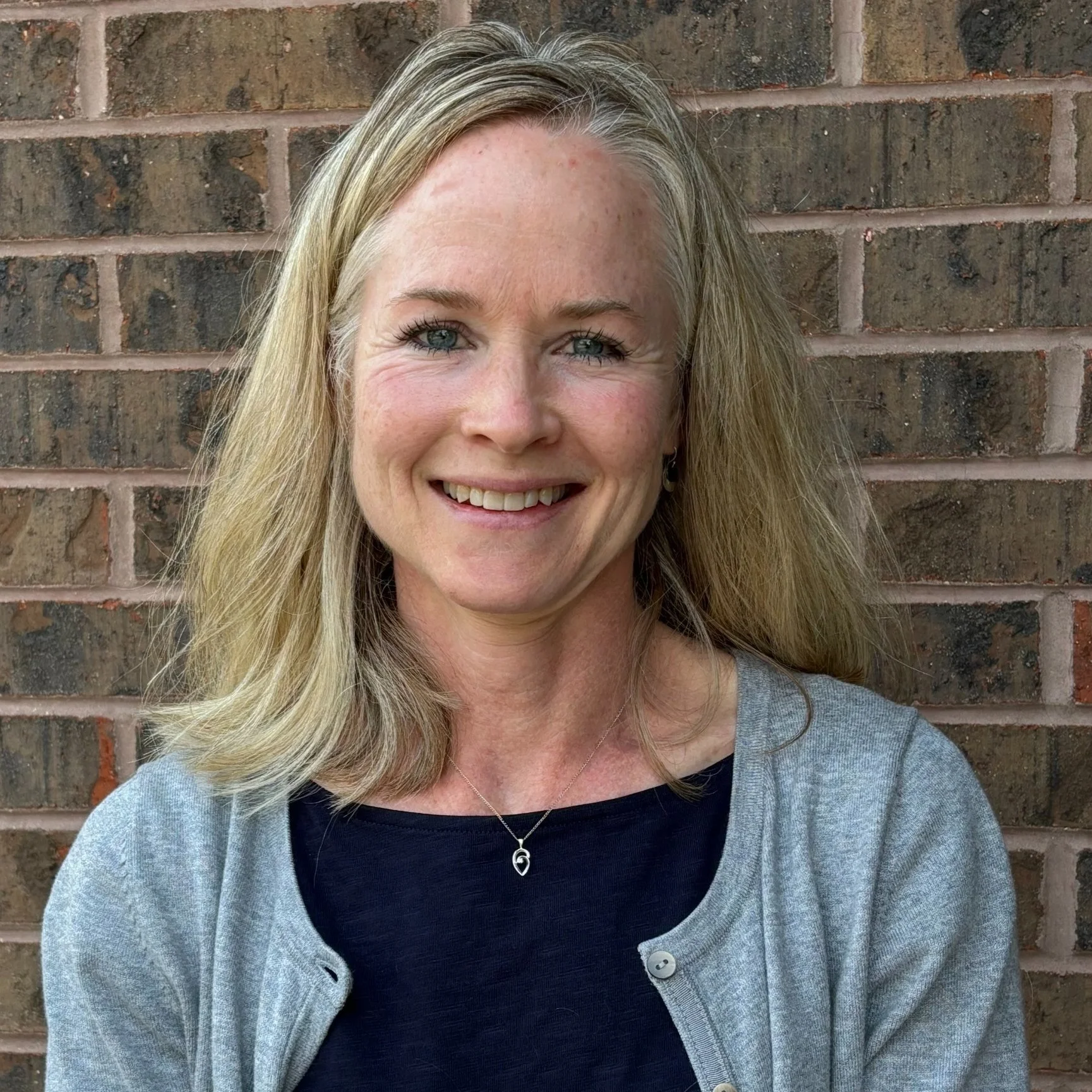 A woman with blonde hair smiling in front of a brick wall, wearing a gray cardigan over a black top and a silver necklace.