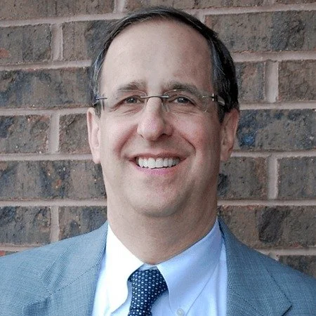 A smiling man in glasses and a light blue suit with a tie, standing in front of a brick wall.
