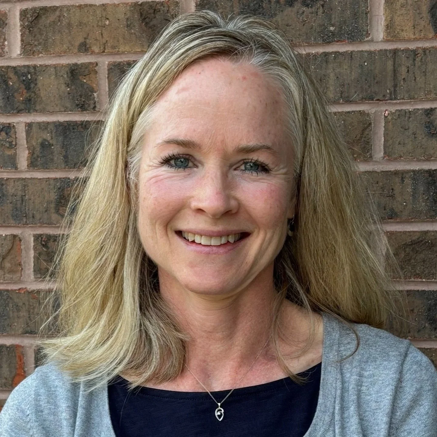 A woman with shoulder-length blonde hair, blue eyes, and fair skin, smiling in front of a brick wall. She is wearing a gray top with a black shirt underneath and a necklace with a pendant.