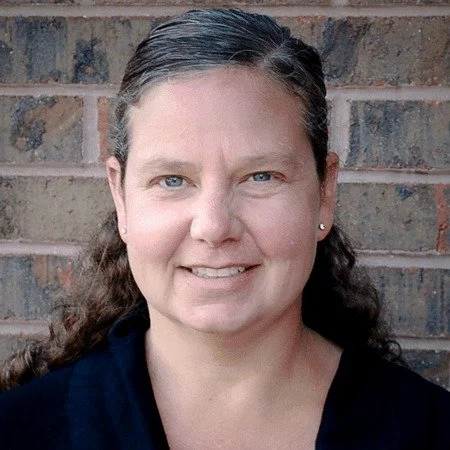 A woman with dark hair, tied back, standing in front of a brick wall, smiling at the camera.