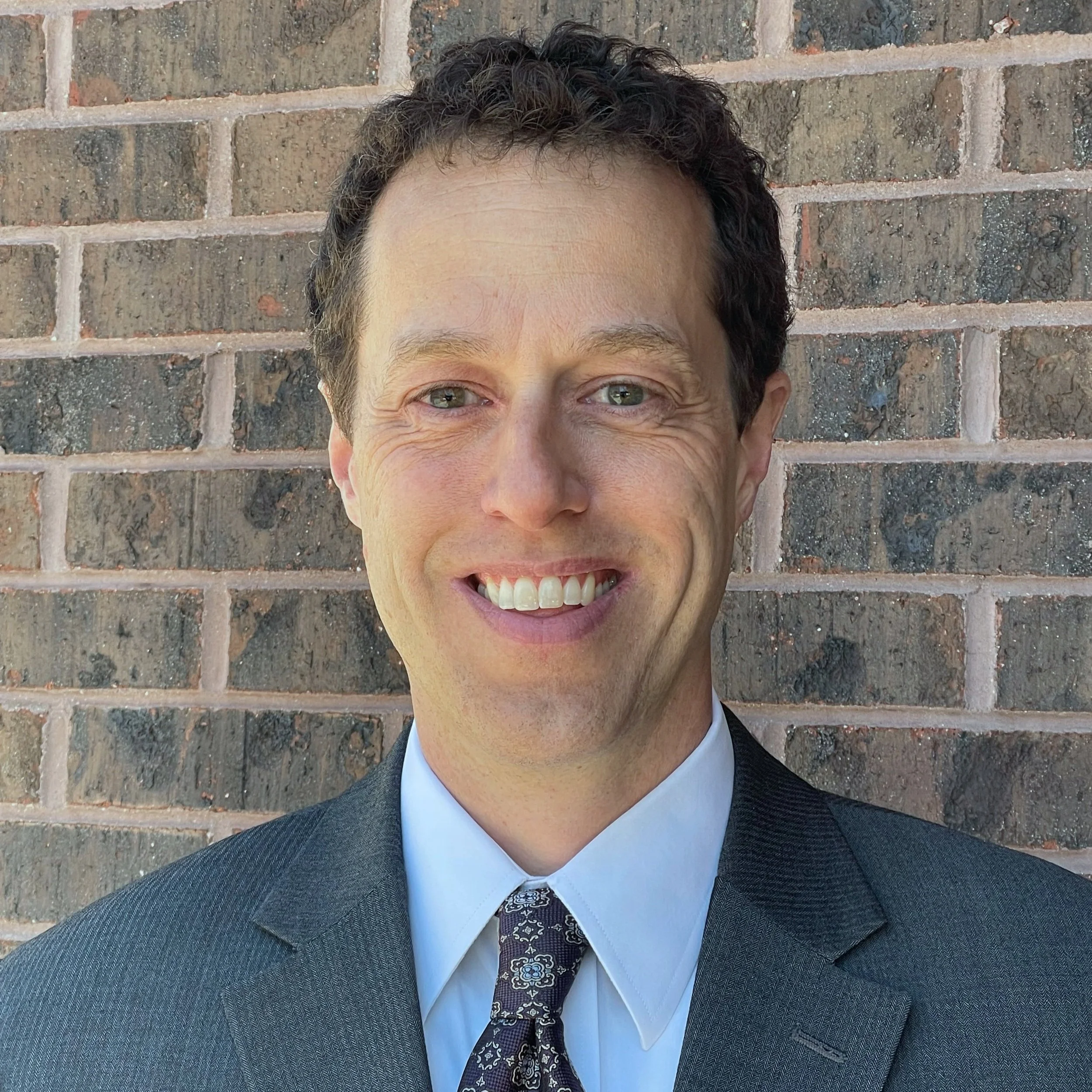A man in a dark suit, light blue shirt, and patterned tie smiling in front of a brick wall.