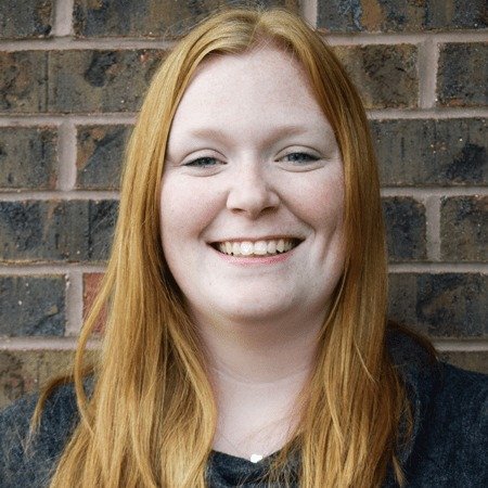 A young woman with long red hair smiling in front of a brick wall.