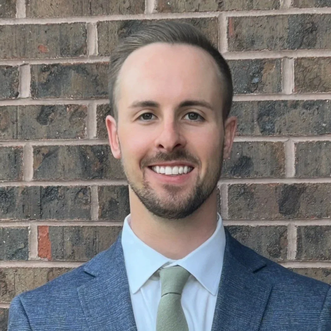 A young man with short brown hair, a beard, and blue eyes, smiling in front of a brick wall. He's wearing a blue suit jacket, a white shirt, and a light green tie.