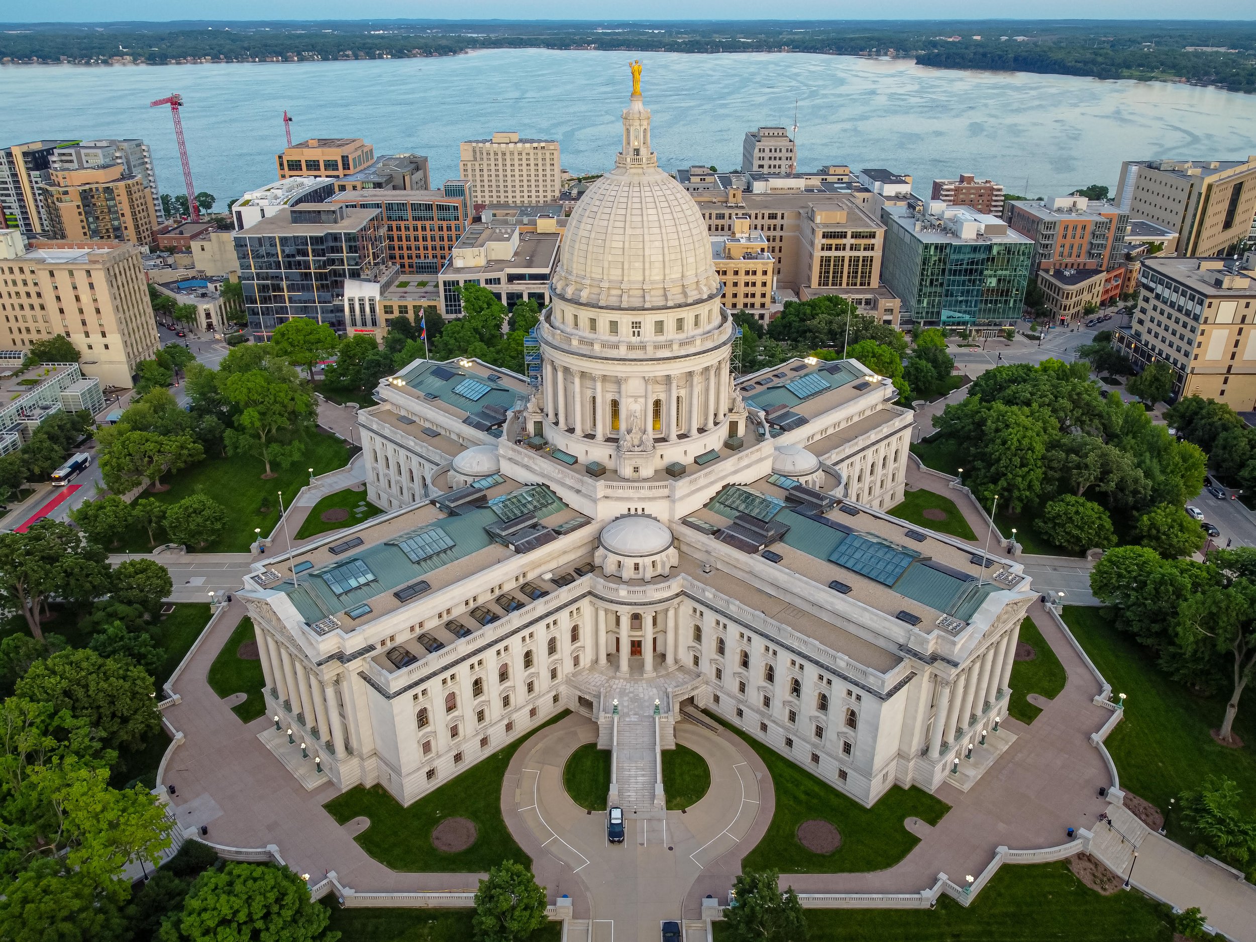 An aerial view of the state capitol building in Sacramento, California. The building features a large dome with a golden statue on top, surrounded by green trees and cityscape buildings, with a river in the background.