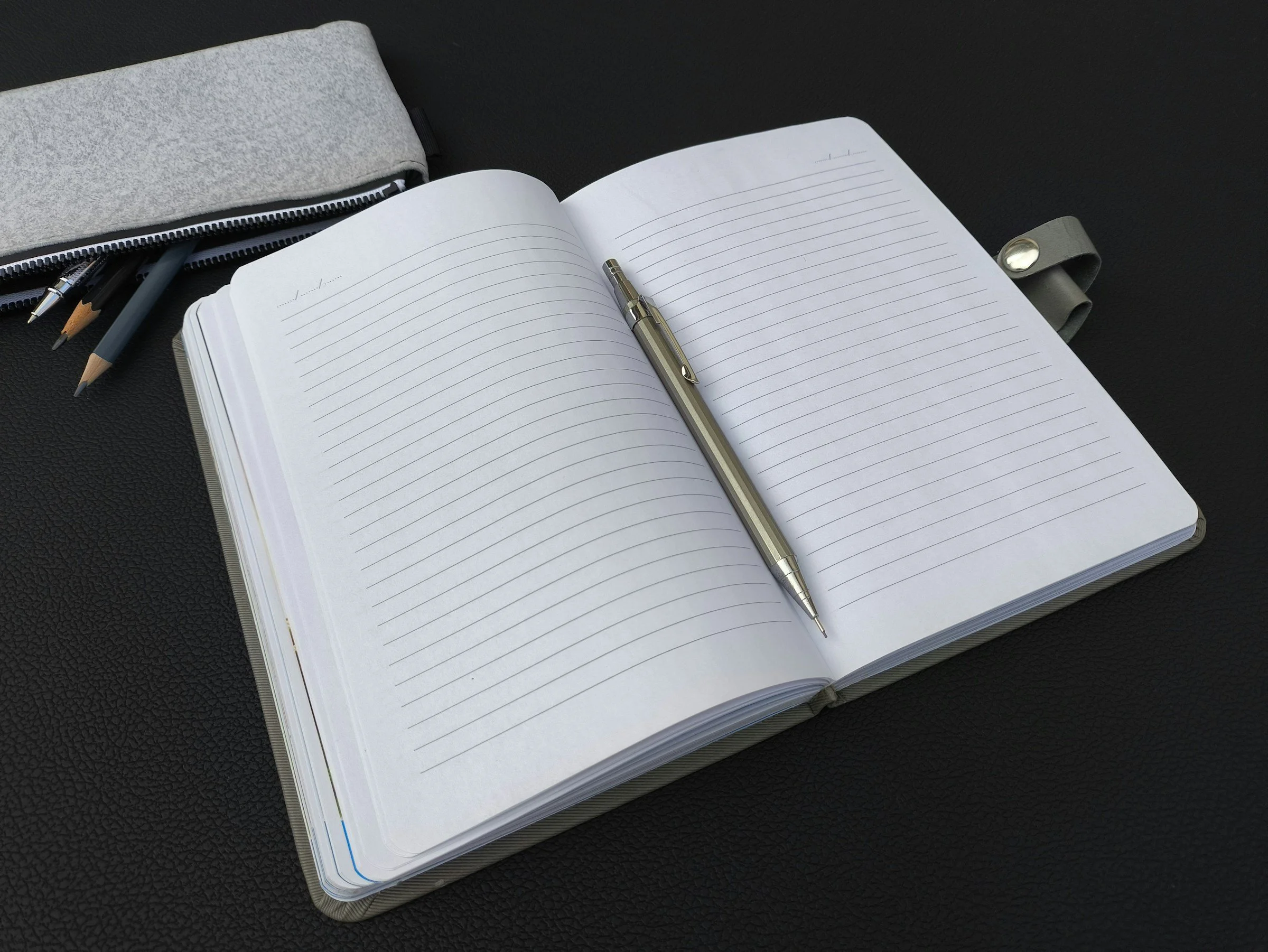 Open blank notebook with lined pages, a silver pen placed in the center, gray pencil case with pens and pencils inside, all on a black textured surface.