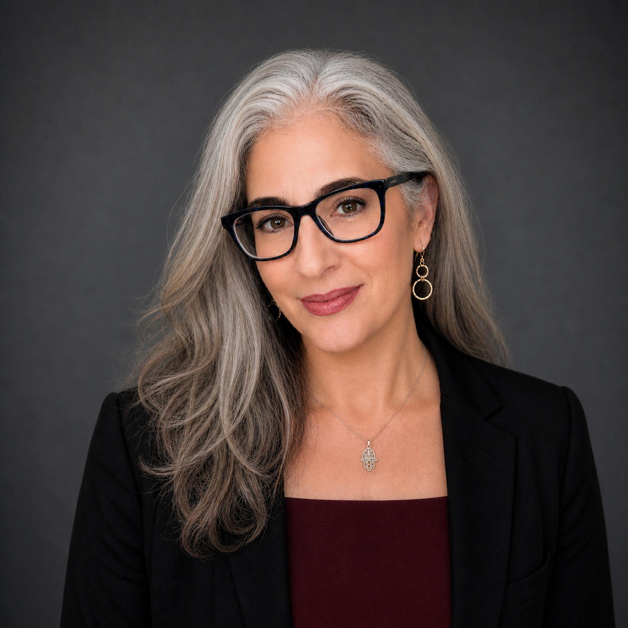 Portrait of a woman with gray hair, wearing glasses, earrings, a necklace, a black blazer, and a burgundy top against a dark gray background.