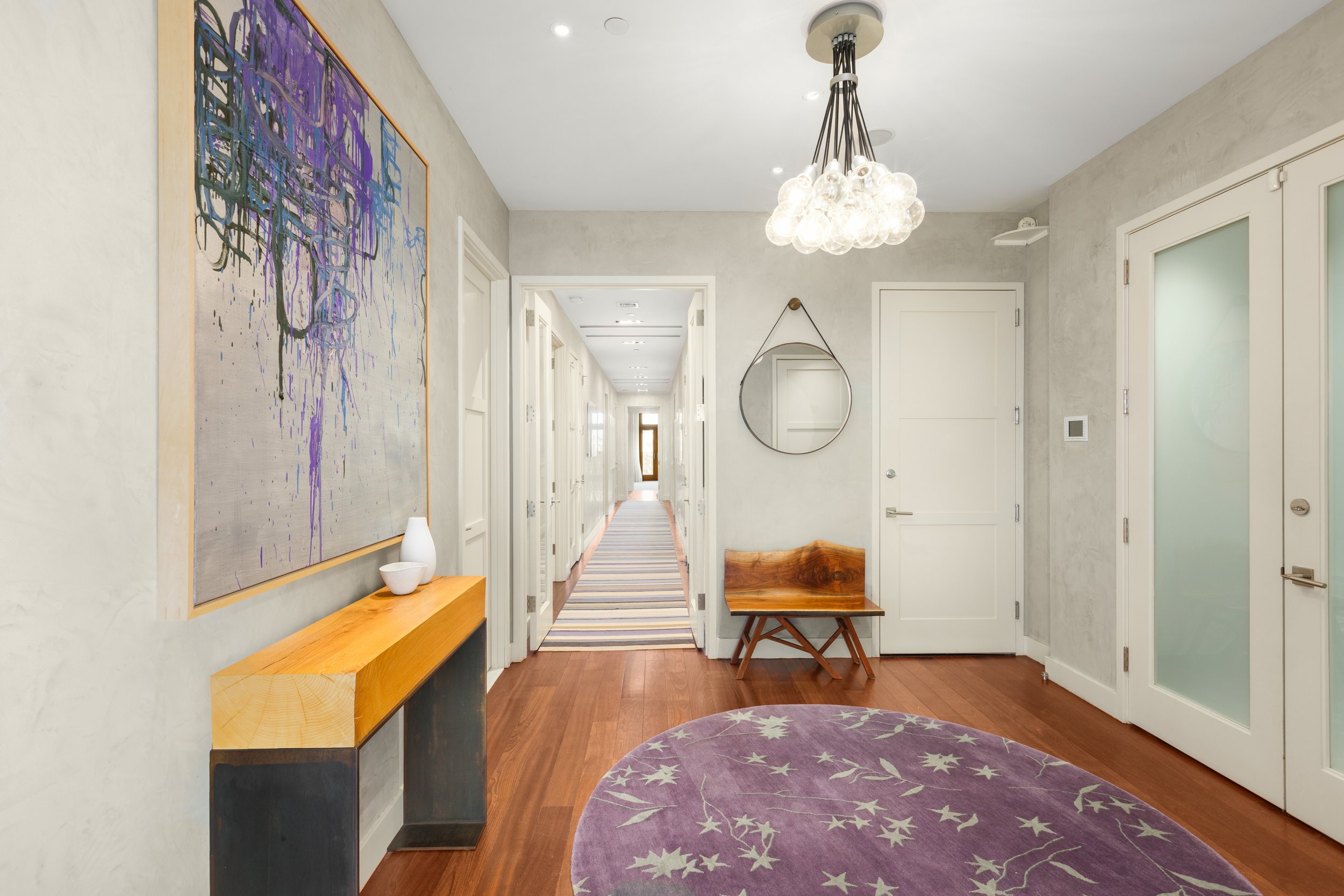 Interior view of a modern hallway with white walls, wooden flooring, and a purple circular rug with star patterns. There is a wooden bench, a round mirror hanging on the wall, a colorful abstract painting, and a ceiling light fixture with multiple gl