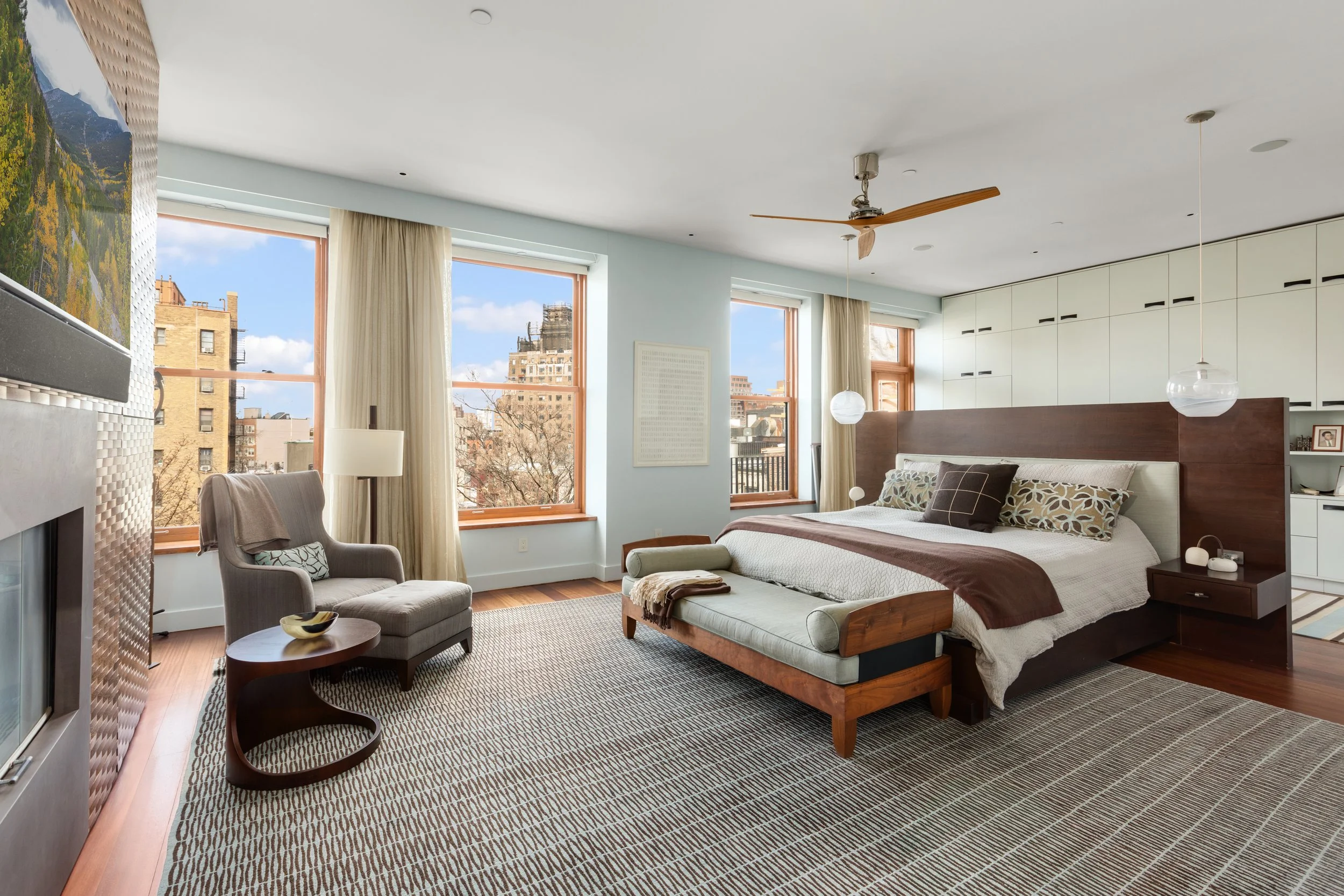 Bright bedroom with large windows showing city buildings, a bed with a wooden headboard, nightstands with lamps, a gray armchair with a blanket, a small side table, a hanging pendant light, and a ceiling fan.