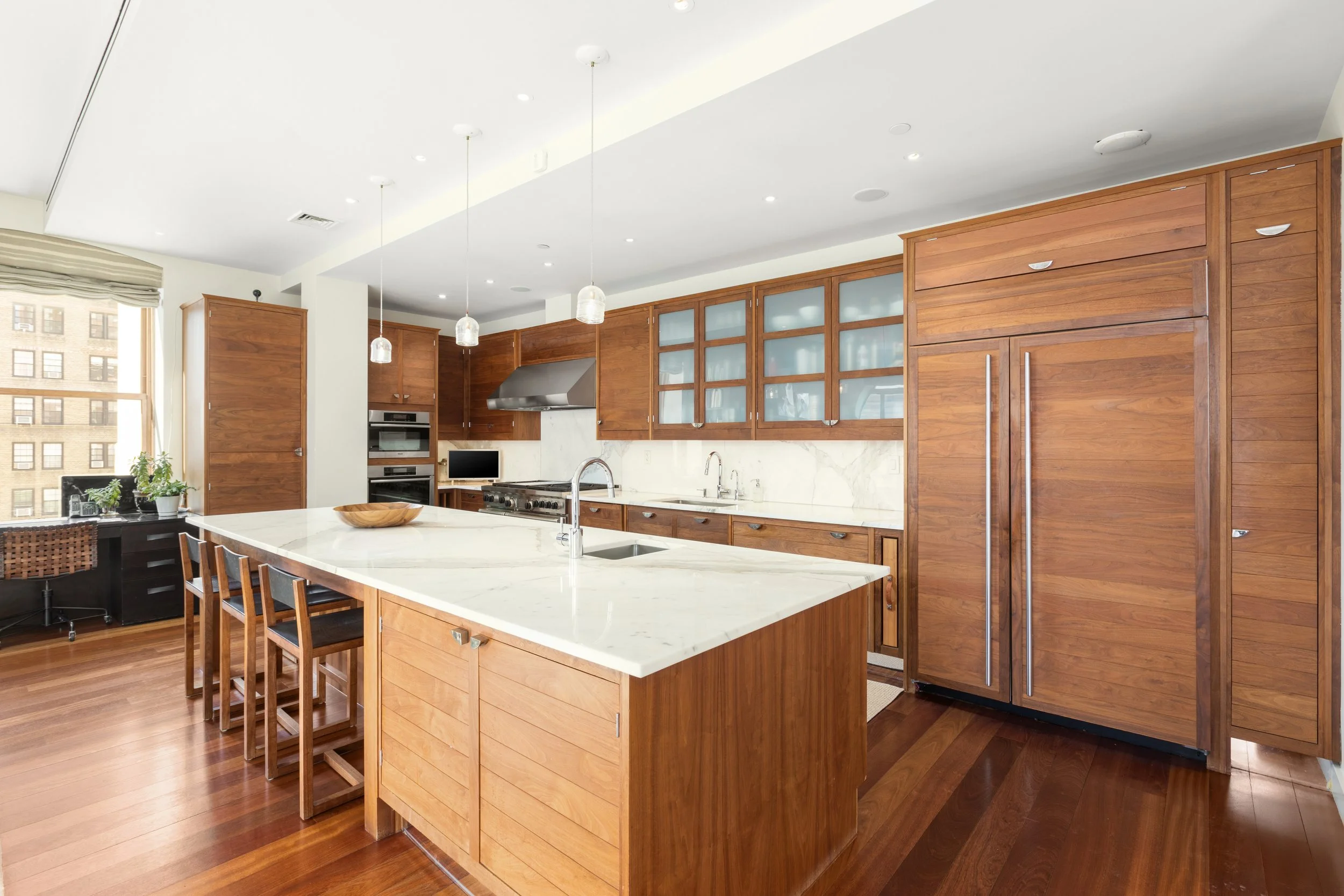 Modern kitchen with wooden cabinetry, white marble countertops, a large island with barstools, and stainless steel appliances, including a refrigerator and oven.
