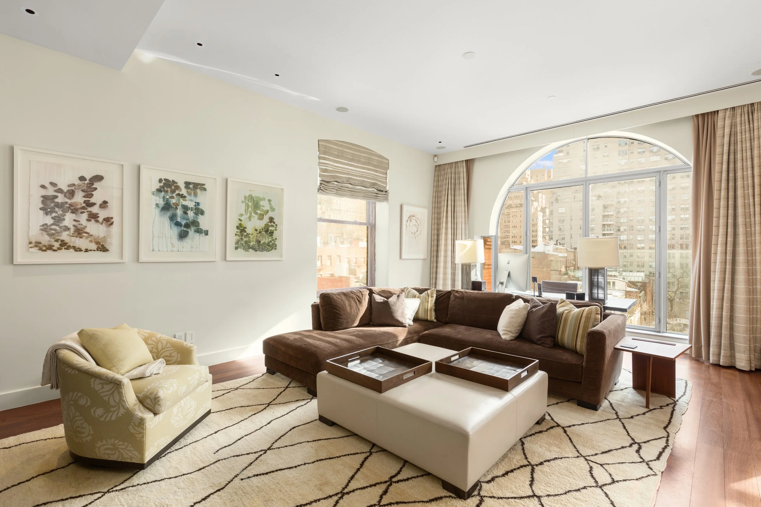 Living room with brown sectional sofa, cream armchair, coffee table with trays, floor-to-ceiling window with city view, beige curtains, decorative pillows, artwork on the wall, and a rug.