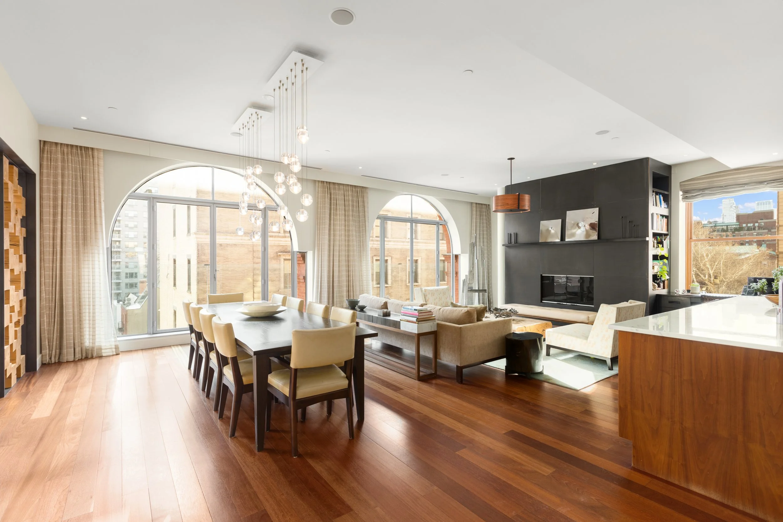 Bright open living and dining room with hardwood floors, large arched windows, beige curtains, modern chandelier, black fireplace, built-in shelves, beige sofa and chairs, coffee table, books, and view of cityscape outside