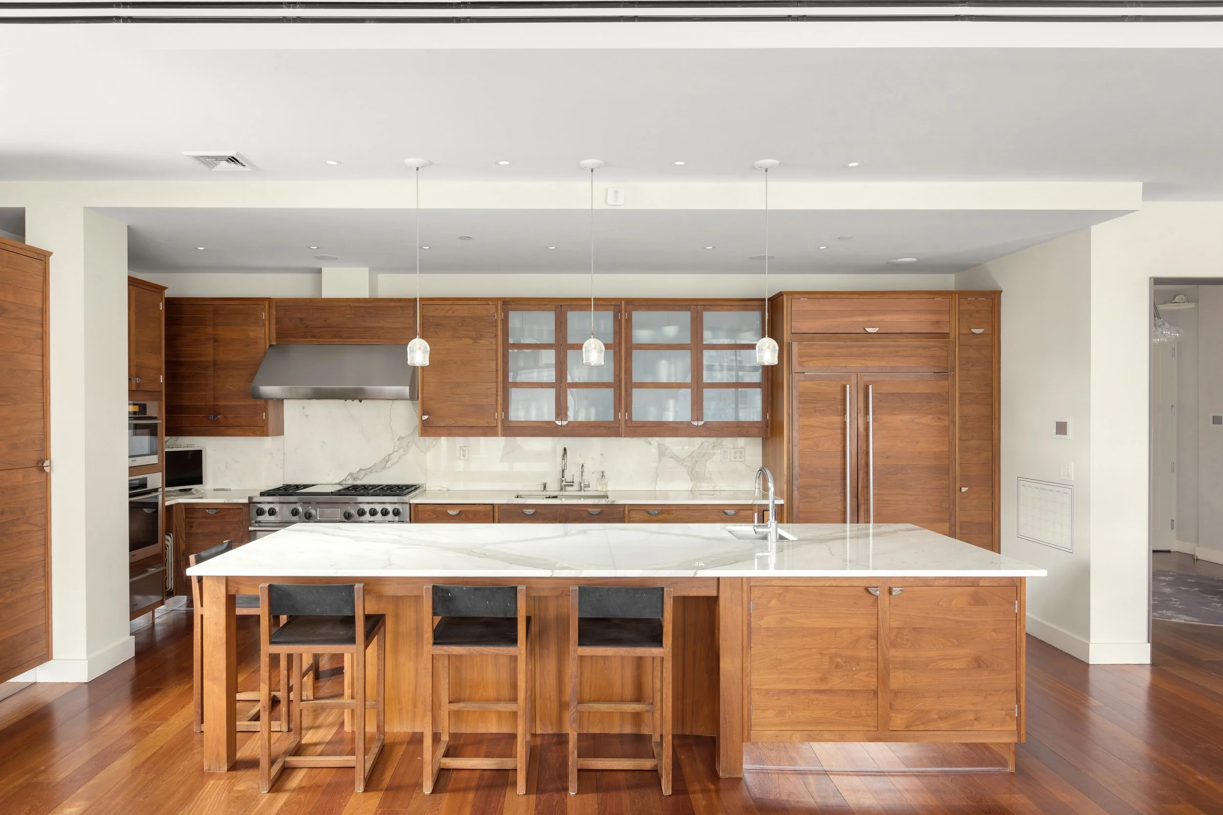 Modern kitchen with wooden cabinetry, marble island, stainless steel appliances, and a hardwood floor.